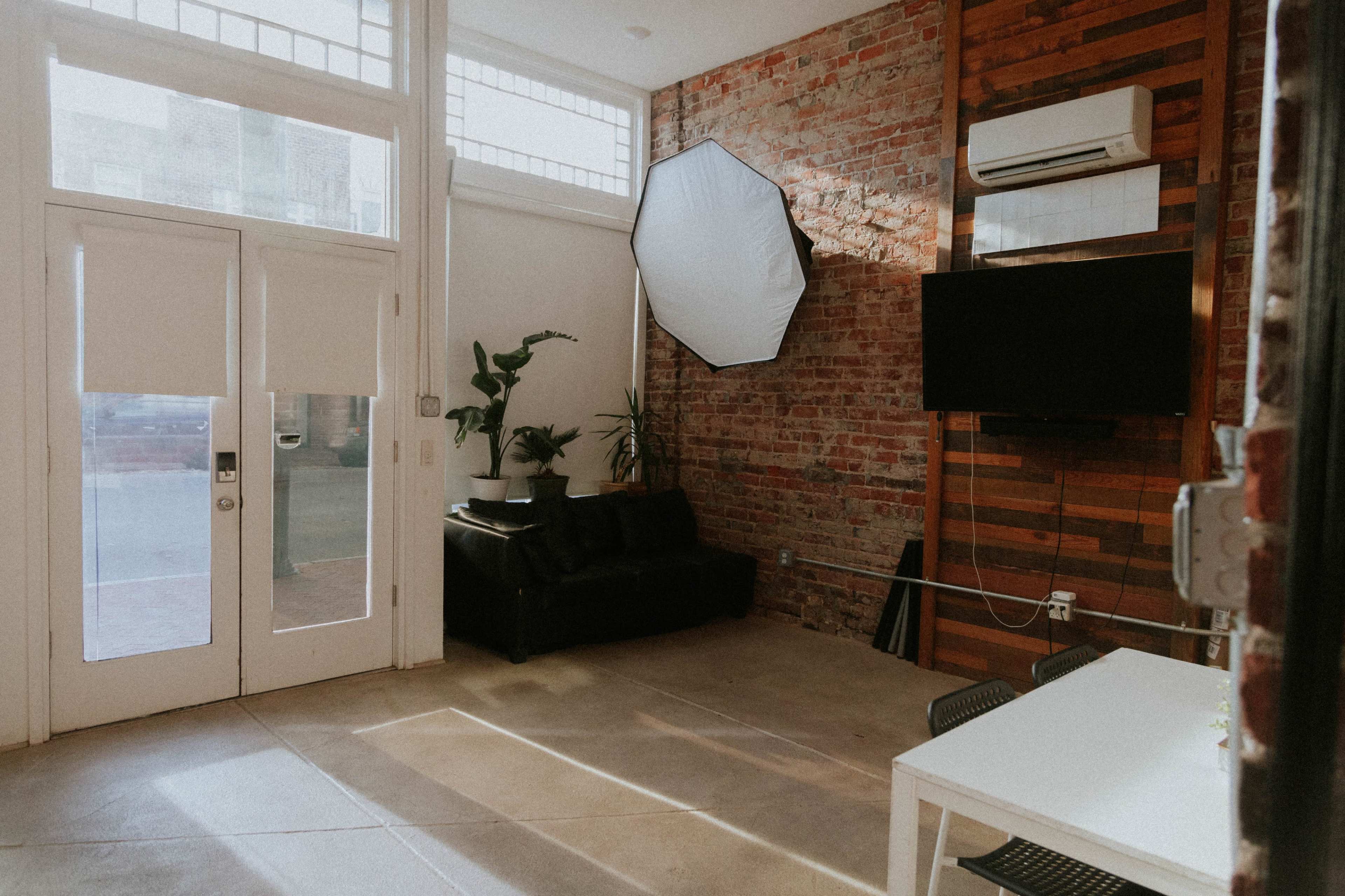 The image shows a modern interior space featuring a black leather sofa, a television mounted on a wooden wall, and a white table with chairs, illuminated by natural light from large windows.