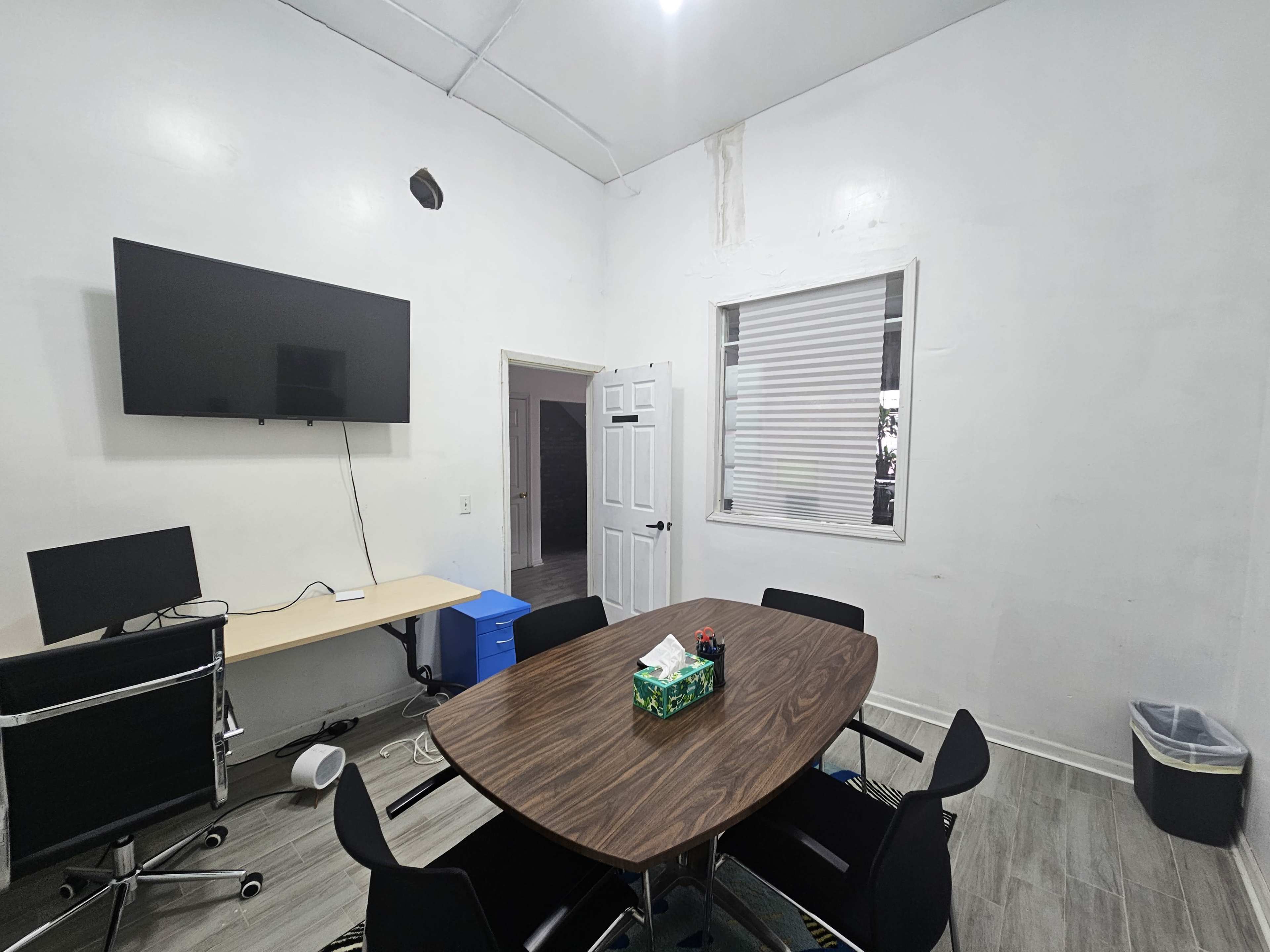 The image shows a small modern meeting room with a large wall-mounted television, a wooden conference table surrounded by black chairs, and a desk with a computer setup.