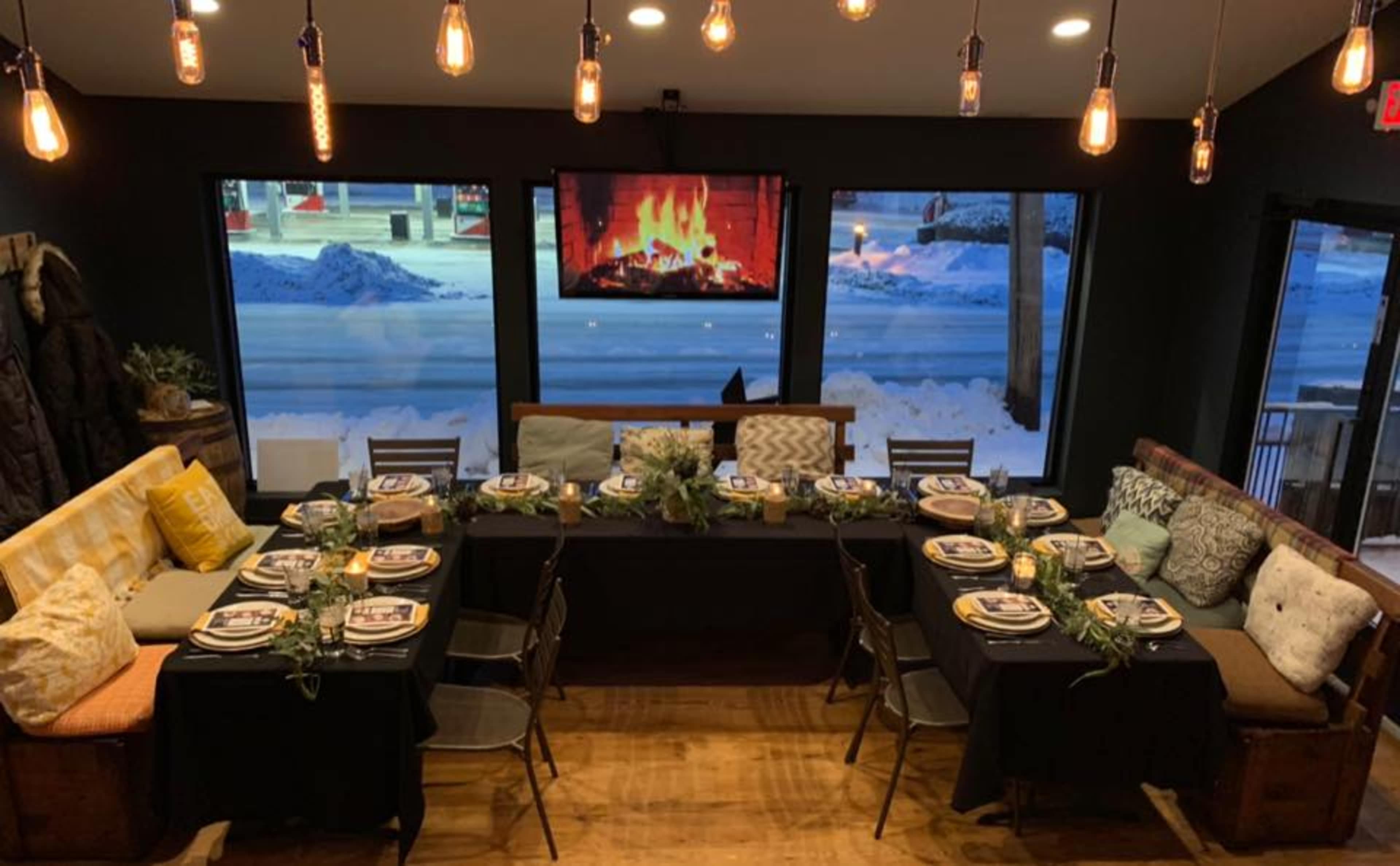 A dining setup features a long table with neatly arranged plates and flowers, surrounded by a cozy seating area and a fireplace displayed on a wall-mounted screen.