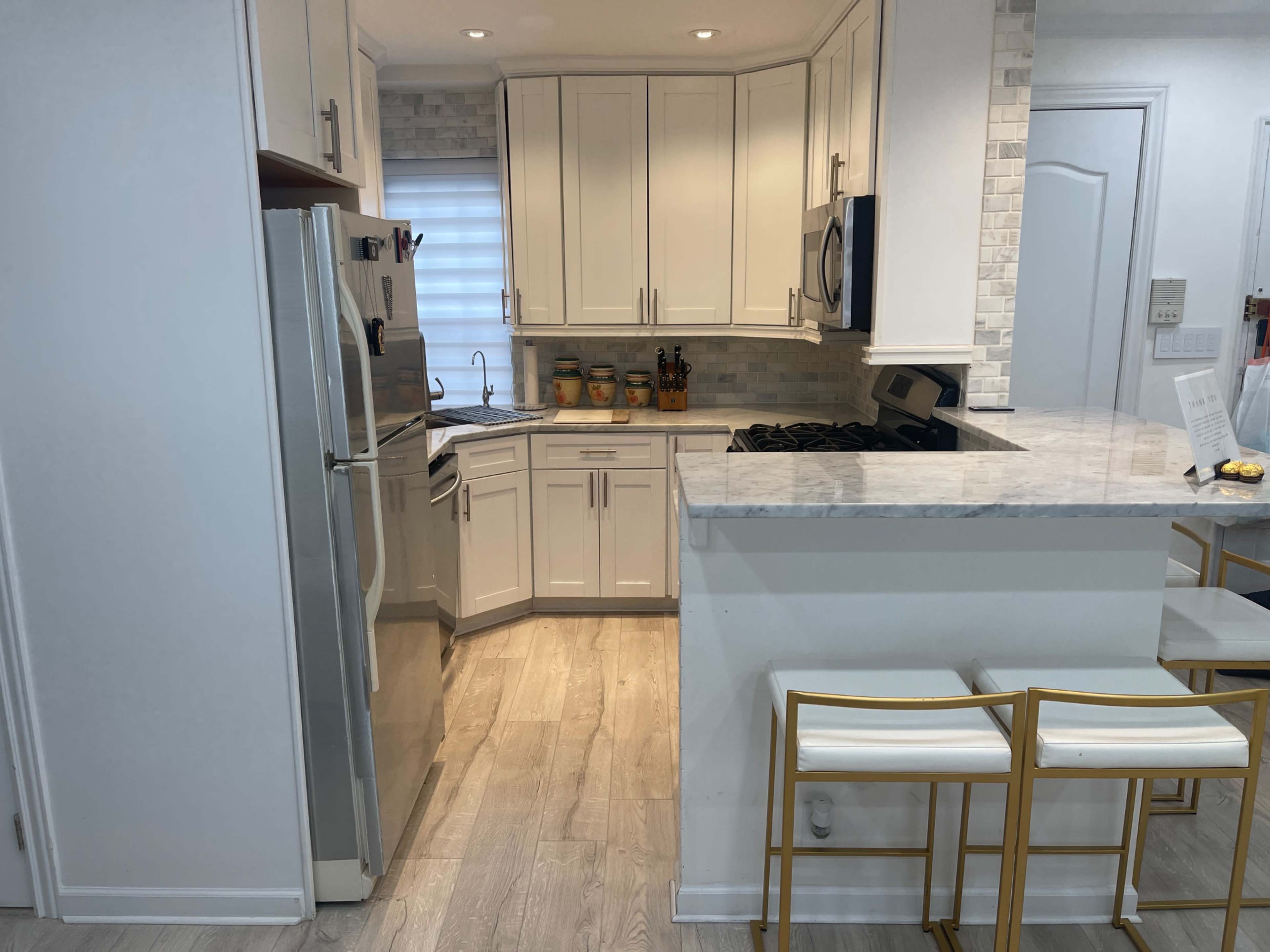 A modern kitchen with white cabinetry, a stainless steel refrigerator, and a marble countertop on a kitchen island with three gold-accented bar stools.