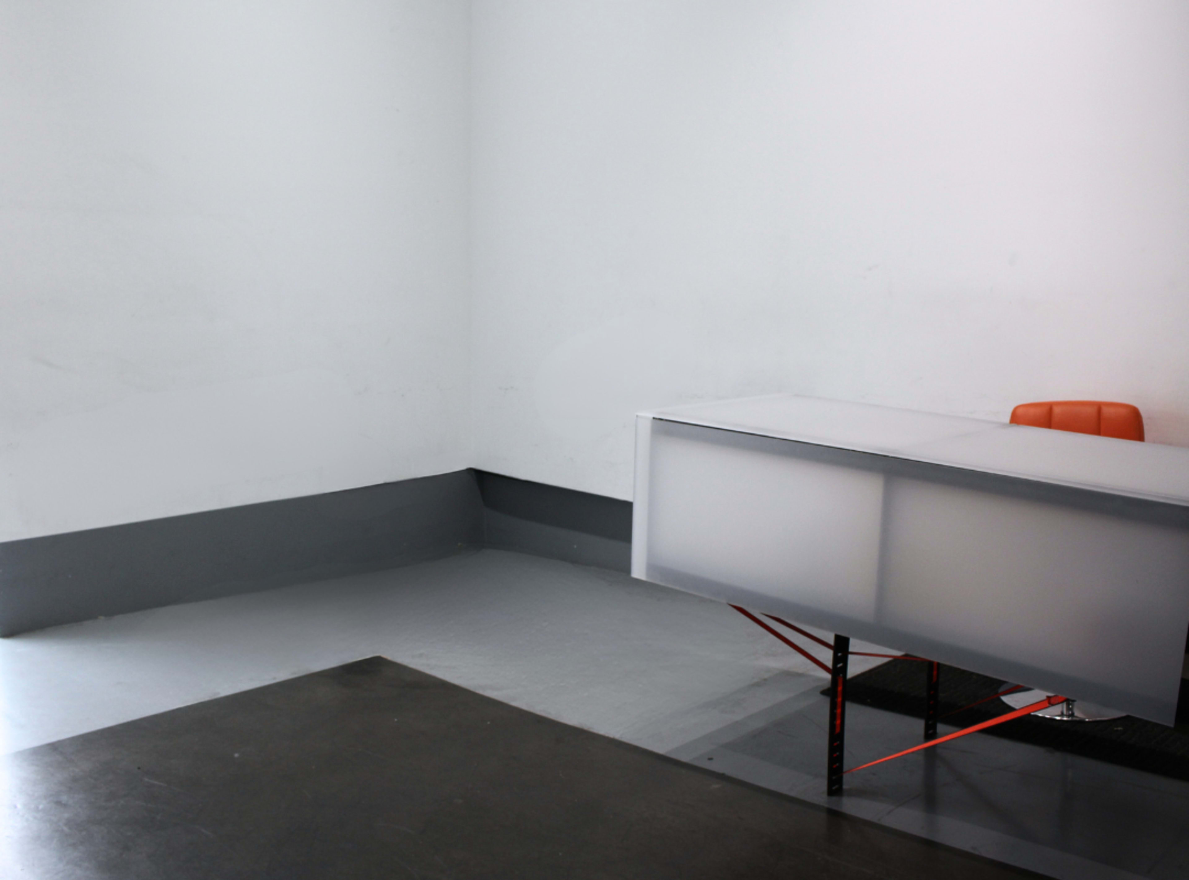 A minimalist office space features a sleek gray floor, a white desk with a frosted front, and a single orange chair.
