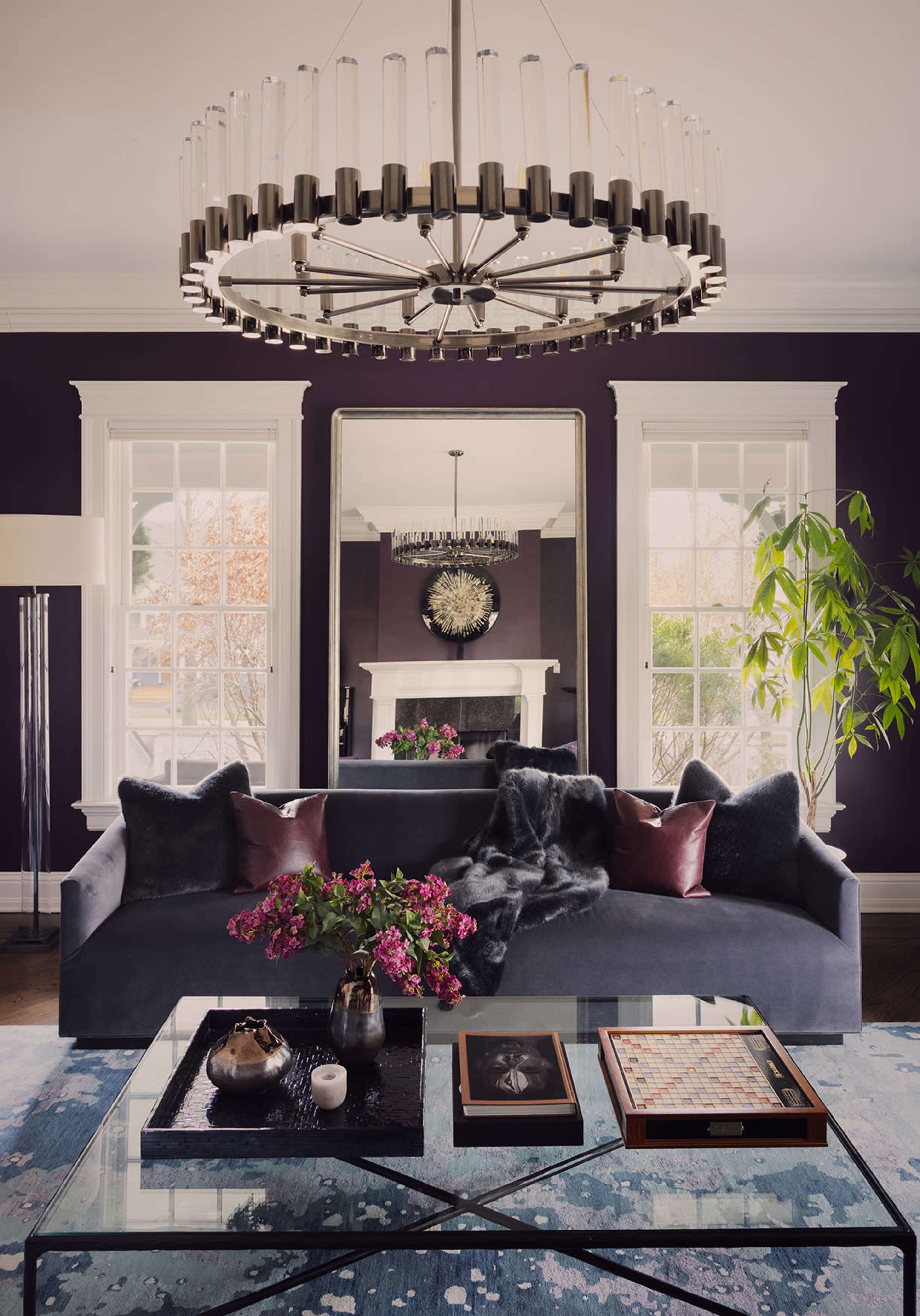 A modern living room features a large chandelier, a dark sofa adorned with pillows, a glass coffee table, and a decorative rug, all set against a deep purple wall and large windows.