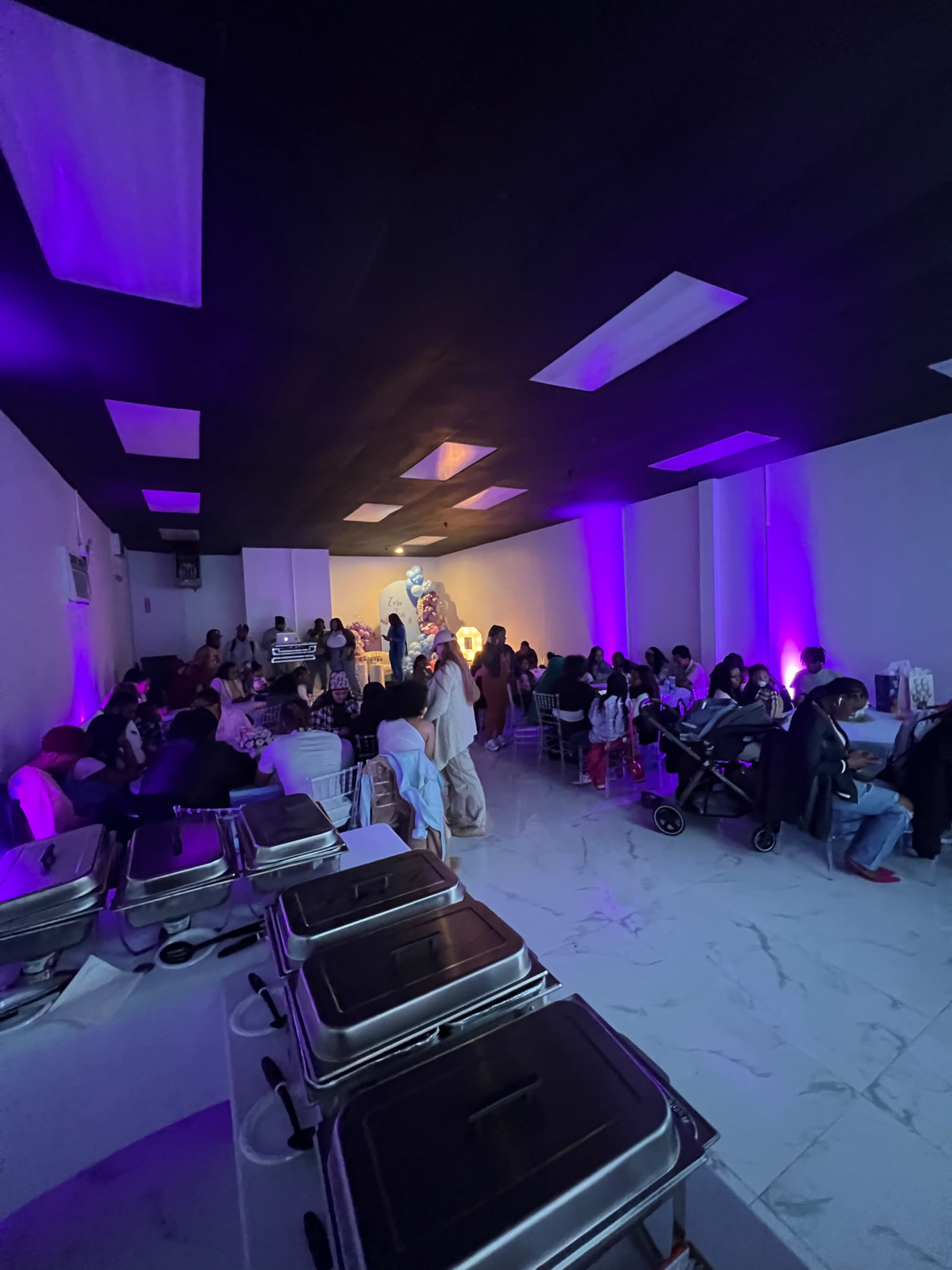 A dimly lit room filled with people seated at tables, some standing, with food trays arranged along the side.