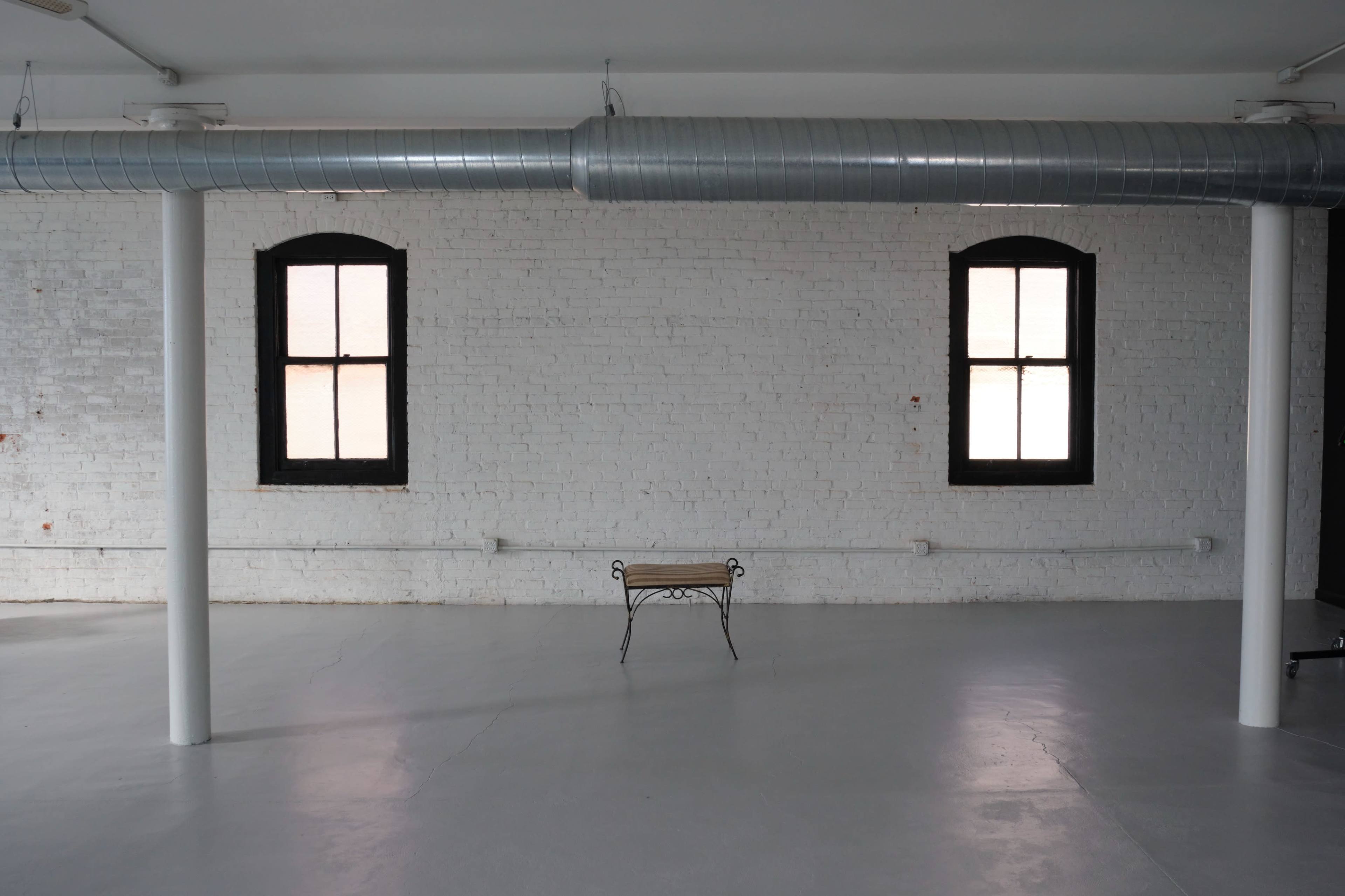 The image shows a vacant room with white brick walls, two windows on opposite sides, and a metal duct running along the ceiling above a small, empty bench in the center.