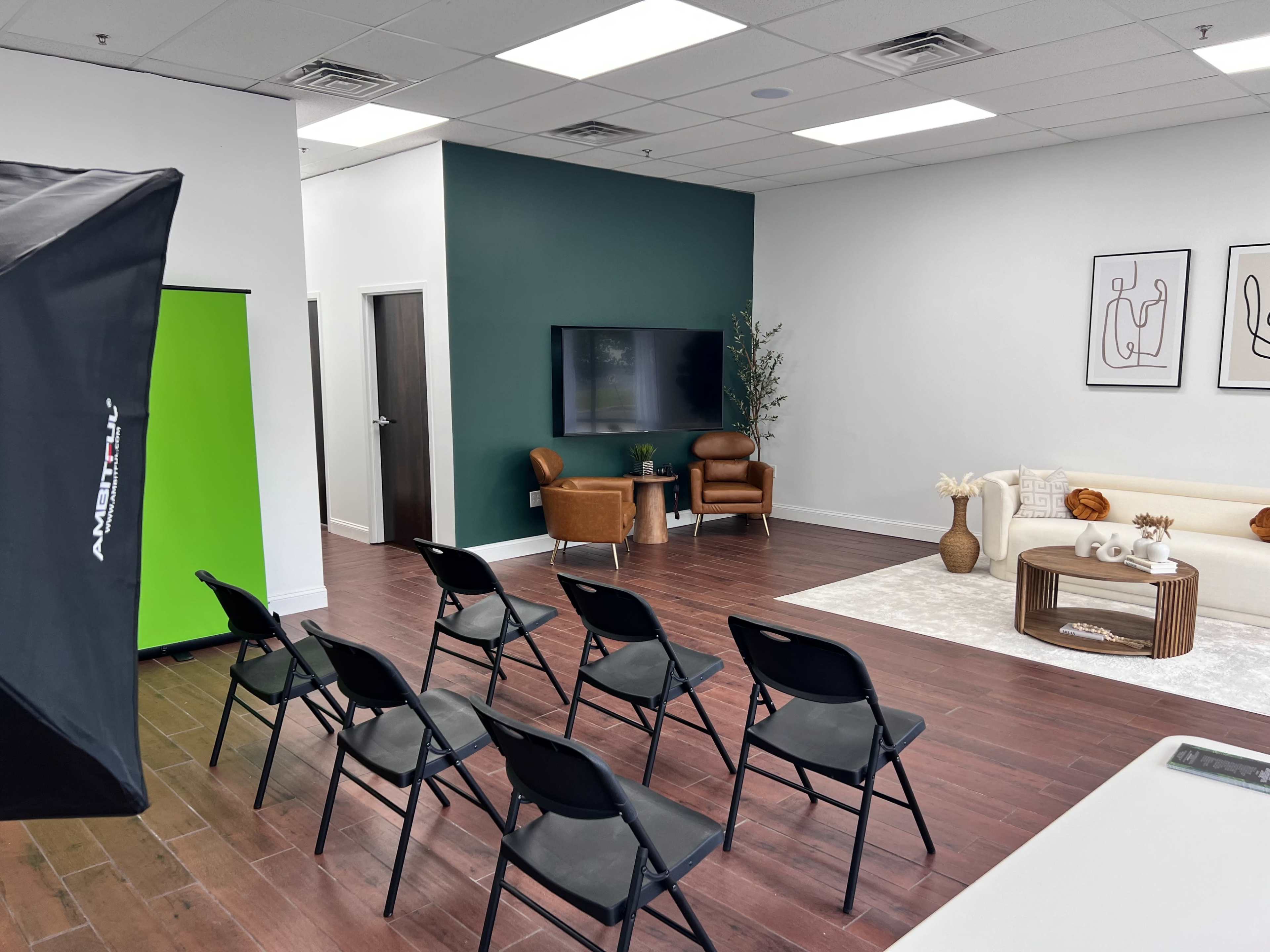 A modern, minimalistic interior with a seating arrangement of black chairs facing a light-colored sofa and a television against a green accent wall.