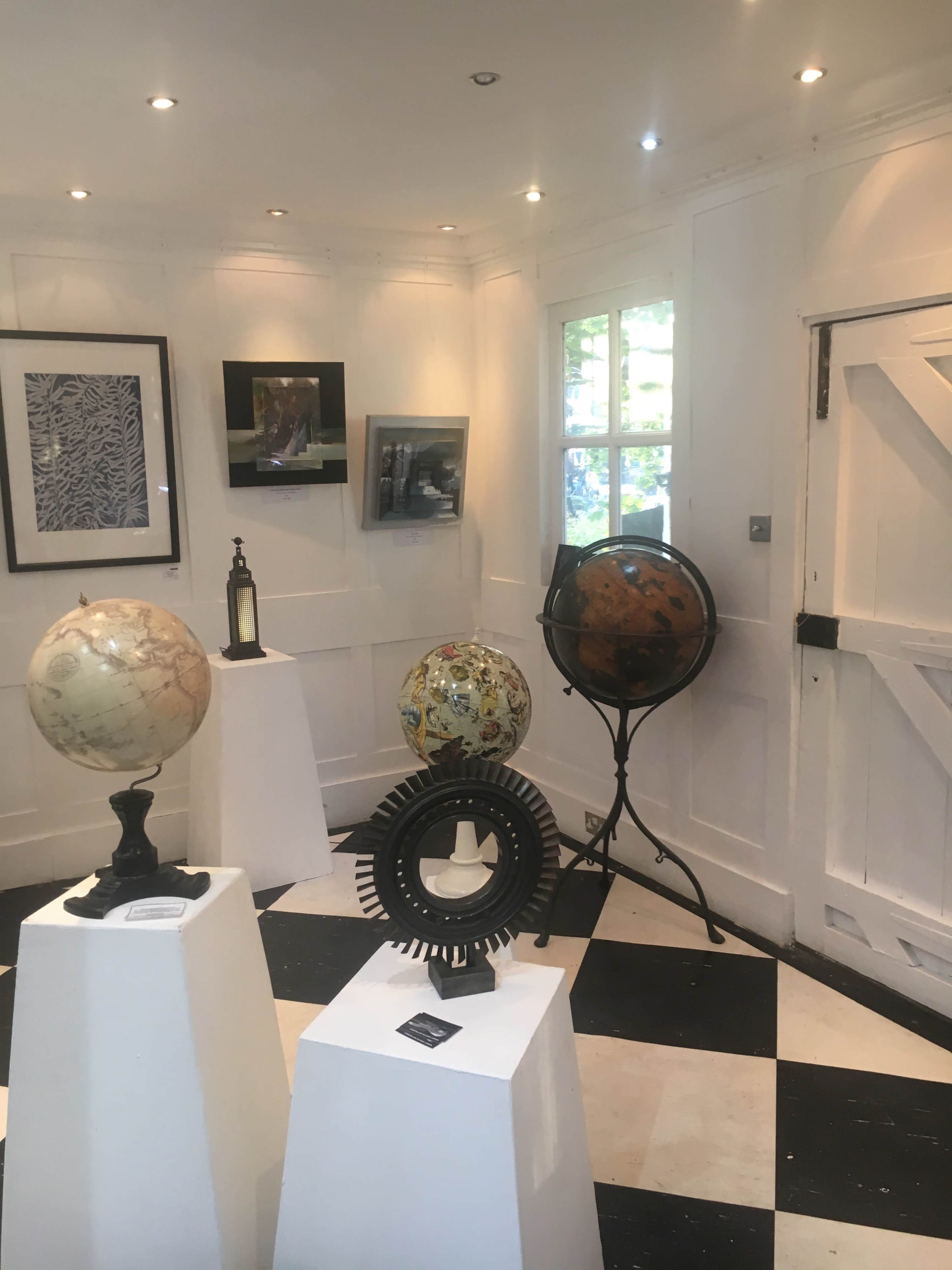 A small art gallery featuring two globes on pedestals, a decorative object resembling a sunburst, and framed artworks on the walls, all under soft lighting.