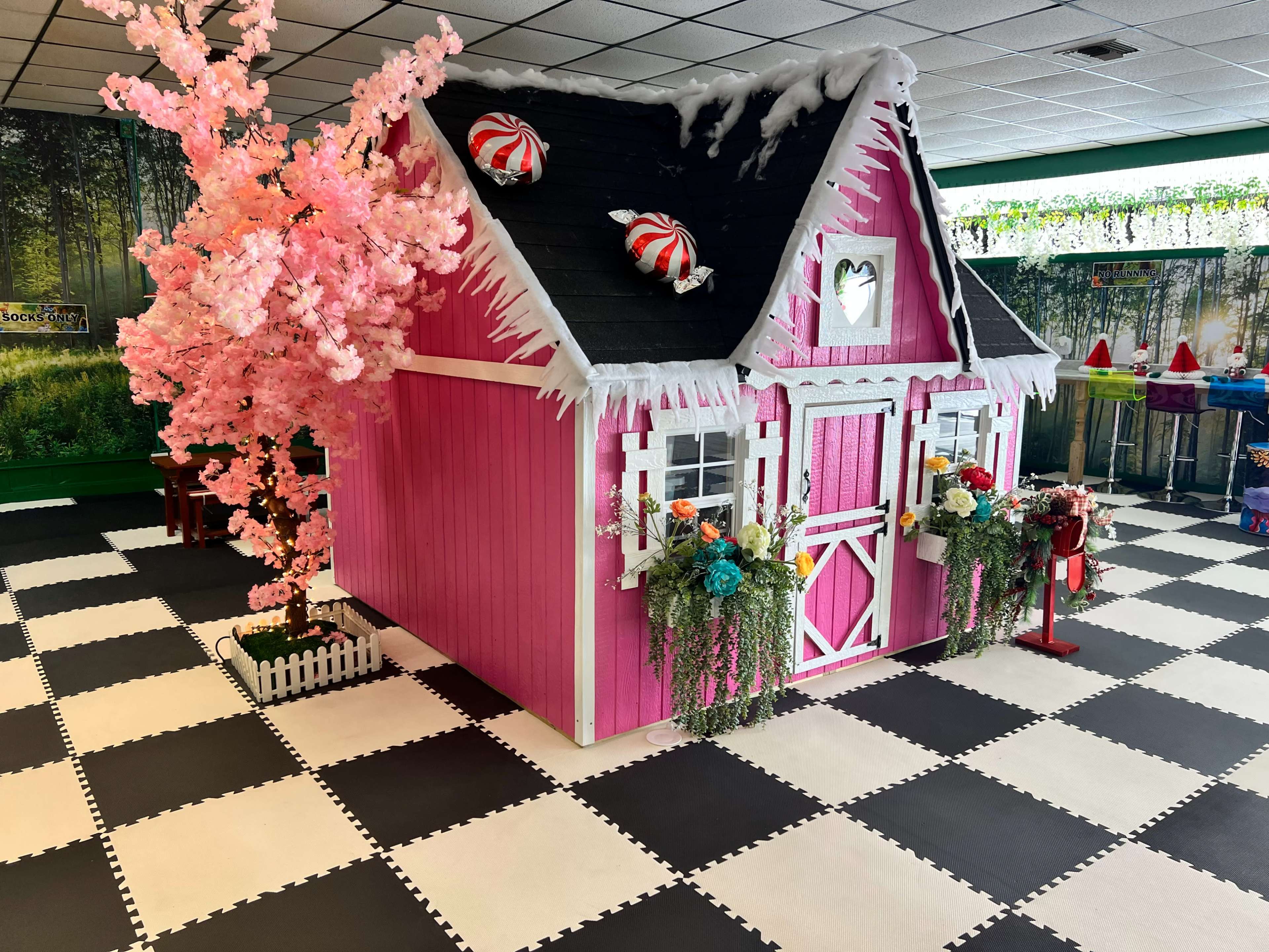 A pink playhouse with decorative elements, including a cherry blossom tree and candy motifs, is set against a black and white checkered floor in a brightly lit room.