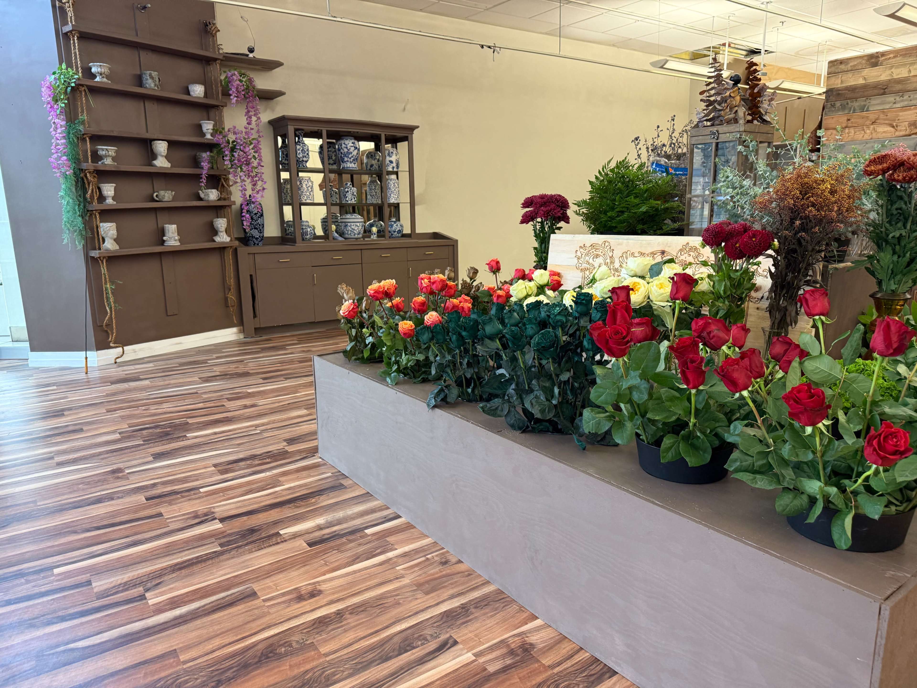 The image shows a floral shop interior with a raised display of potted red and yellow flowers, a wooden floor, and a decorated wall featuring shelves and furniture.