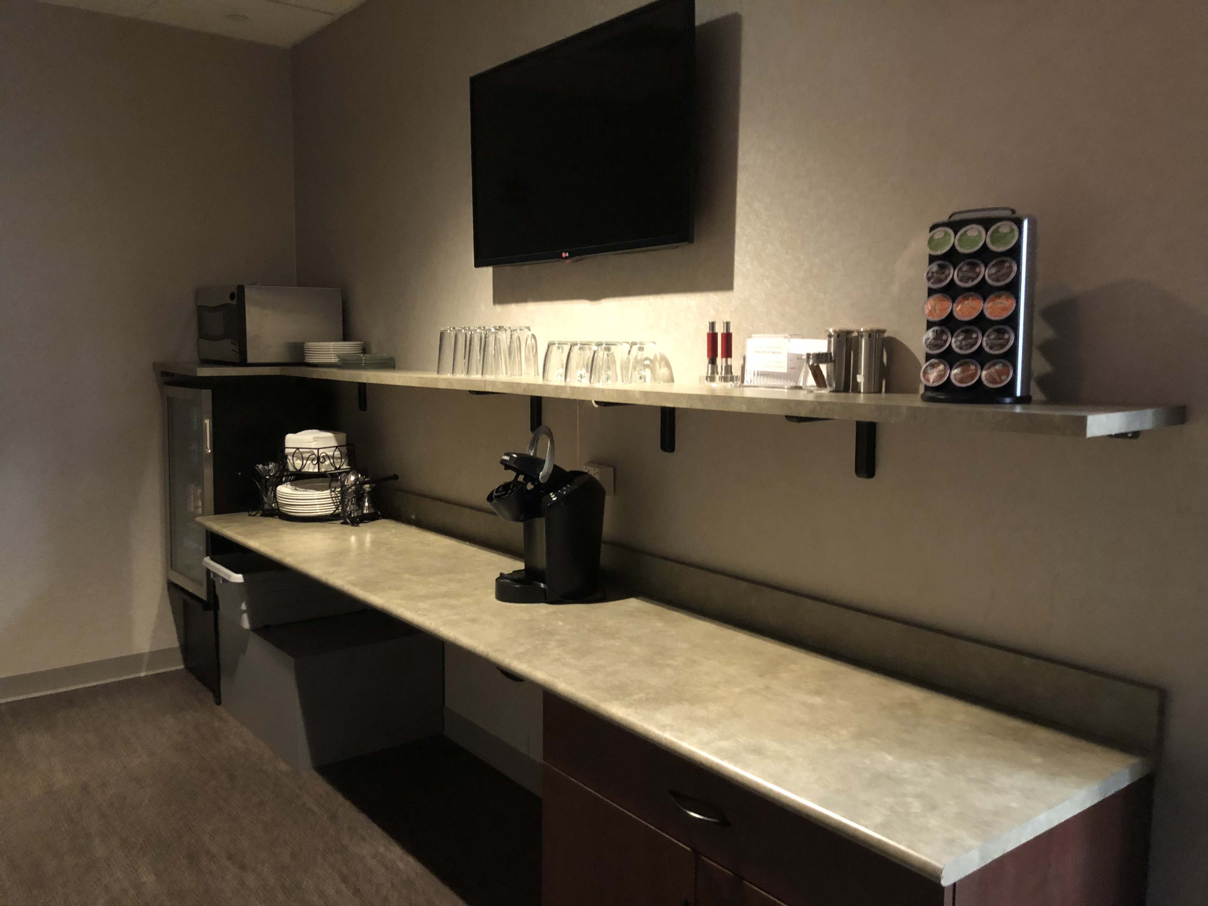 The image shows a small kitchenette area featuring a countertop with a coffee maker, a selection of coffee pods, glasses, and a refrigerator underneath.