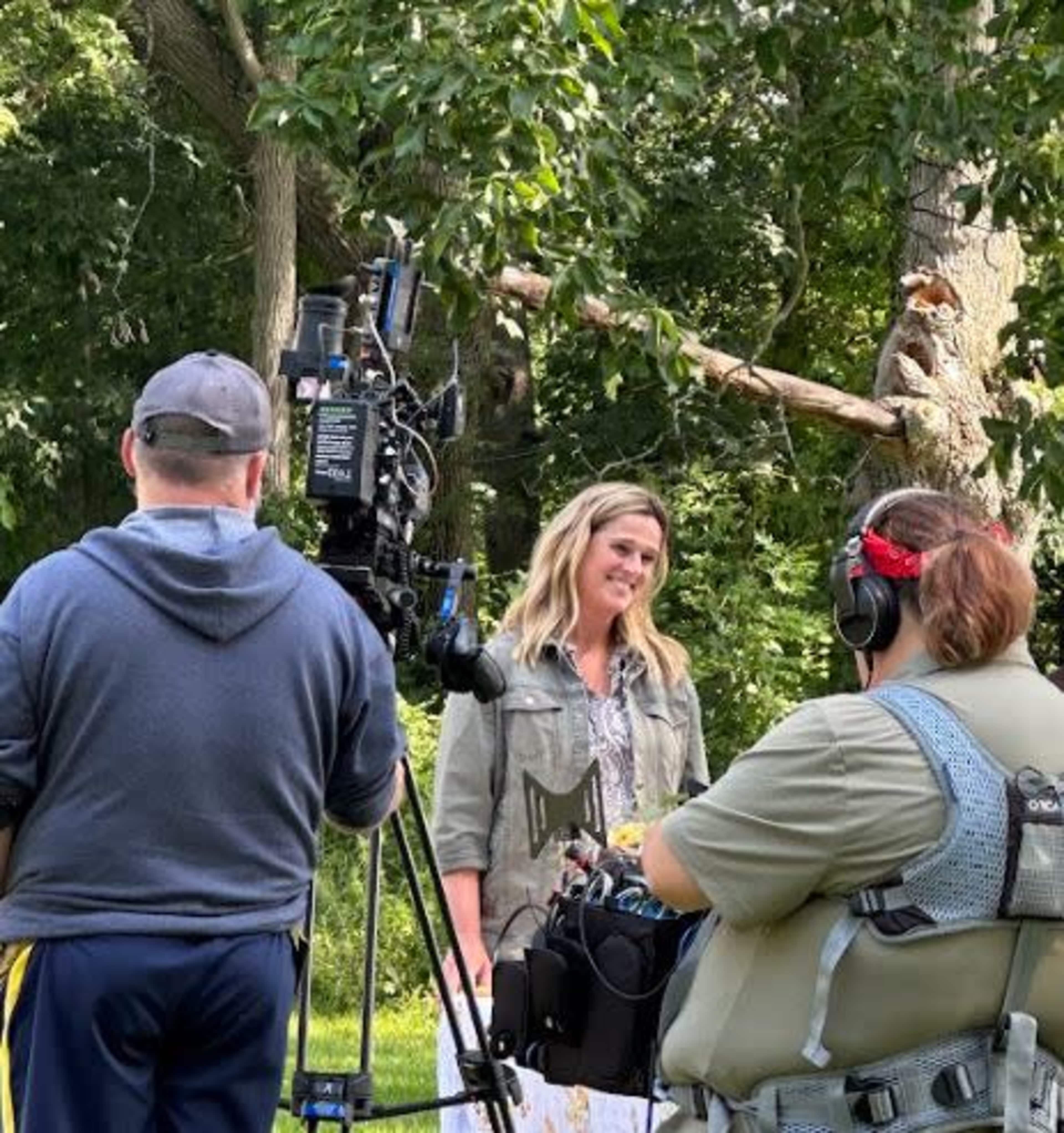 A film crew sets up in a wooded area as a woman stands smiling in front of the camera.