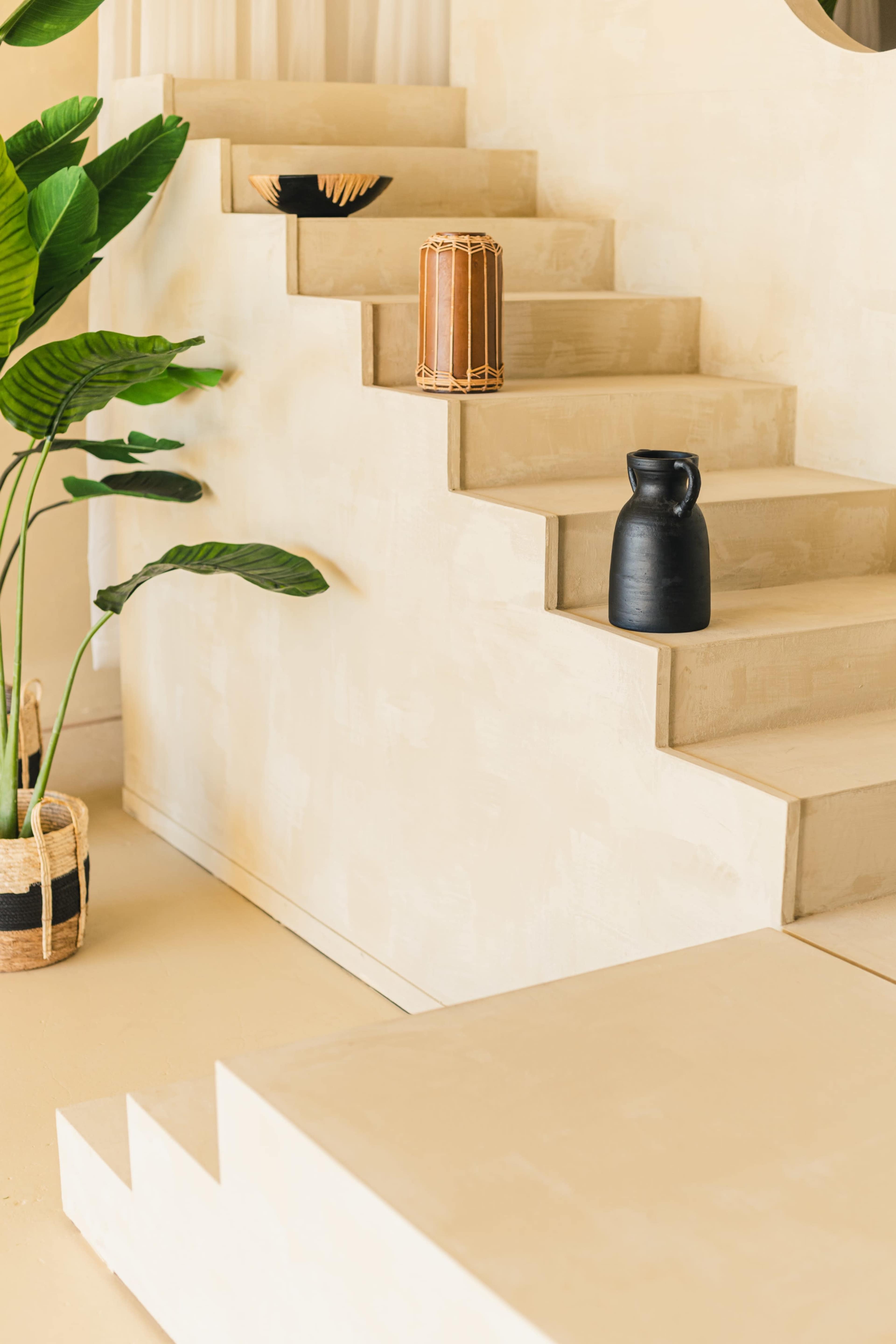 A staircase with textured beige steps, adorned with a black vase, a cylindrical wooden drum, and a decorative woven basket near a large potted plant.