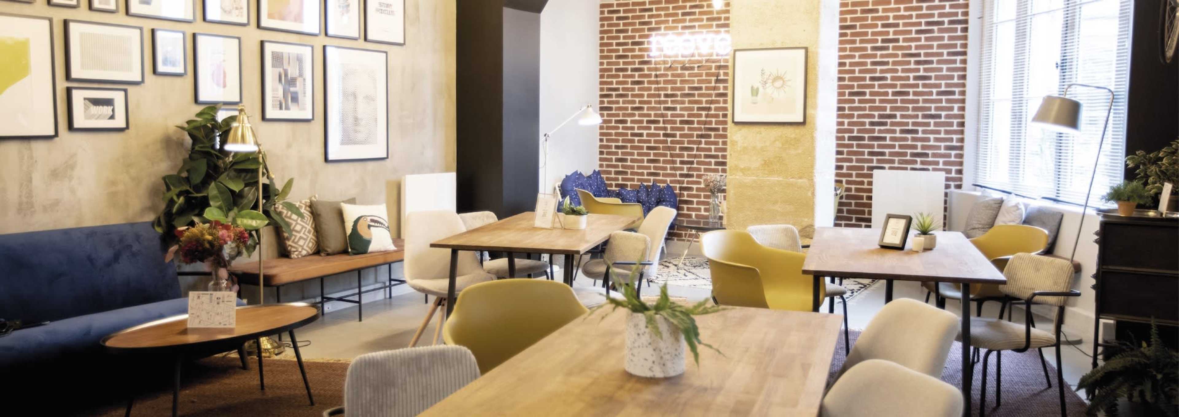 A cozy interior space features wooden tables surrounded by various seating arrangements, framed artwork on the walls, and plants accentuating the decor.