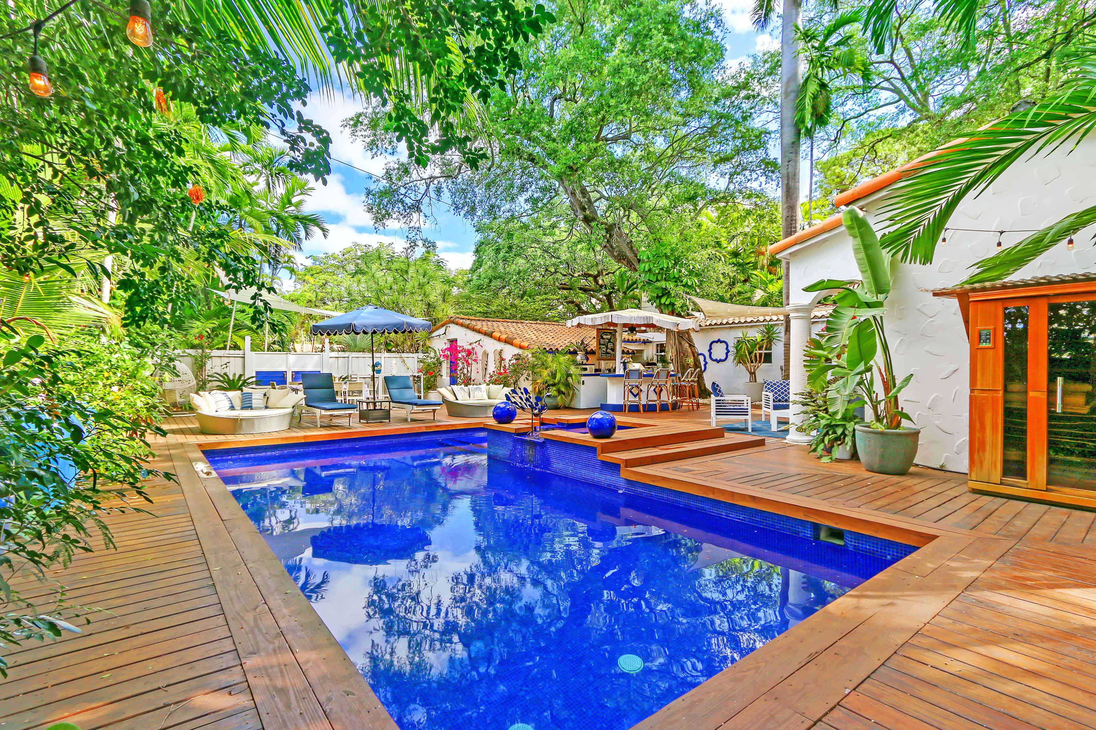 A rectangular swimming pool surrounded by wooden decking, lounge chairs, and lush greenery, with a patio area and various outdoor furnishings nearby.