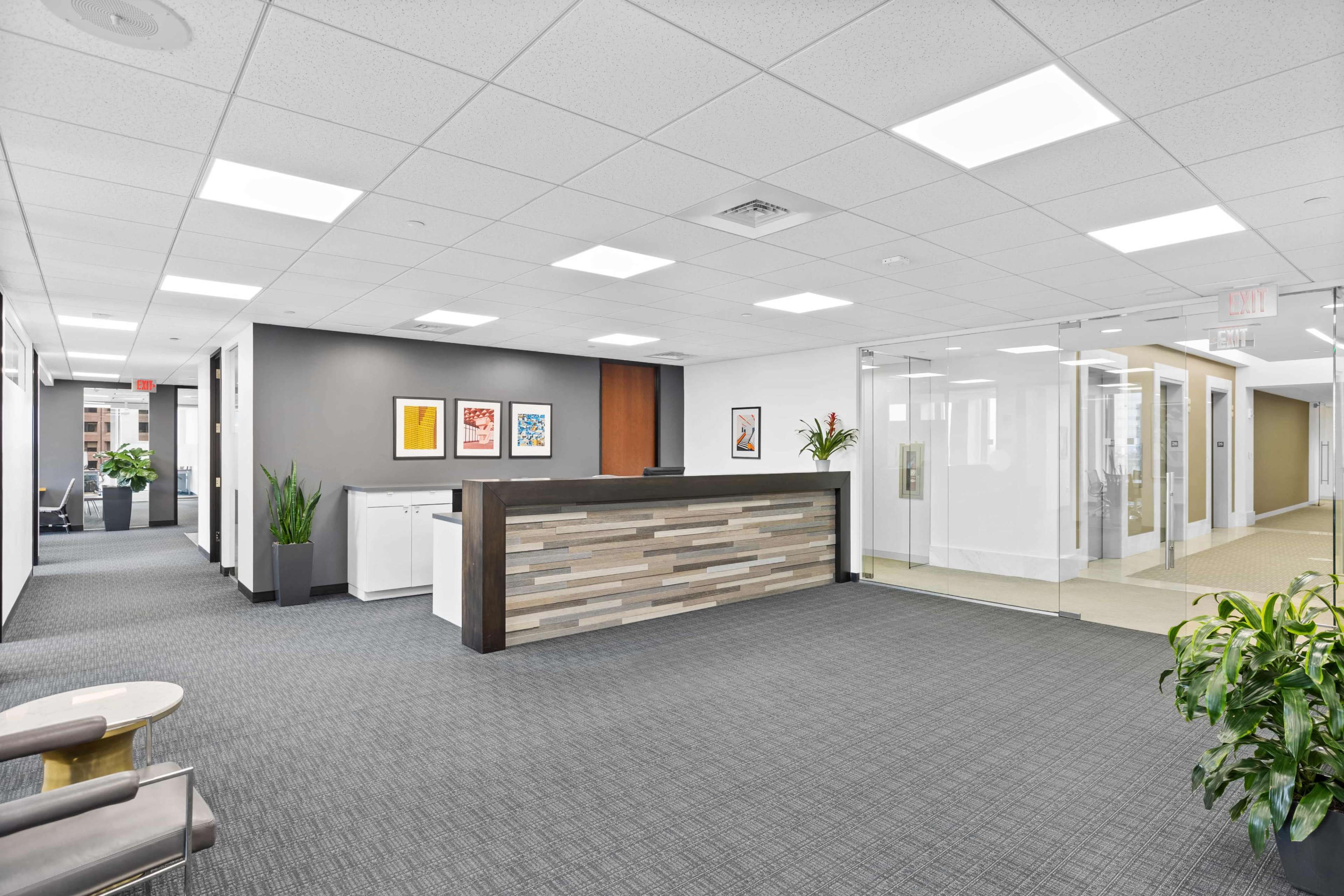 The image shows a modern office reception area featuring a wooden reception desk, contemporary artwork on the walls, and a clean, well-lit environment with plants.