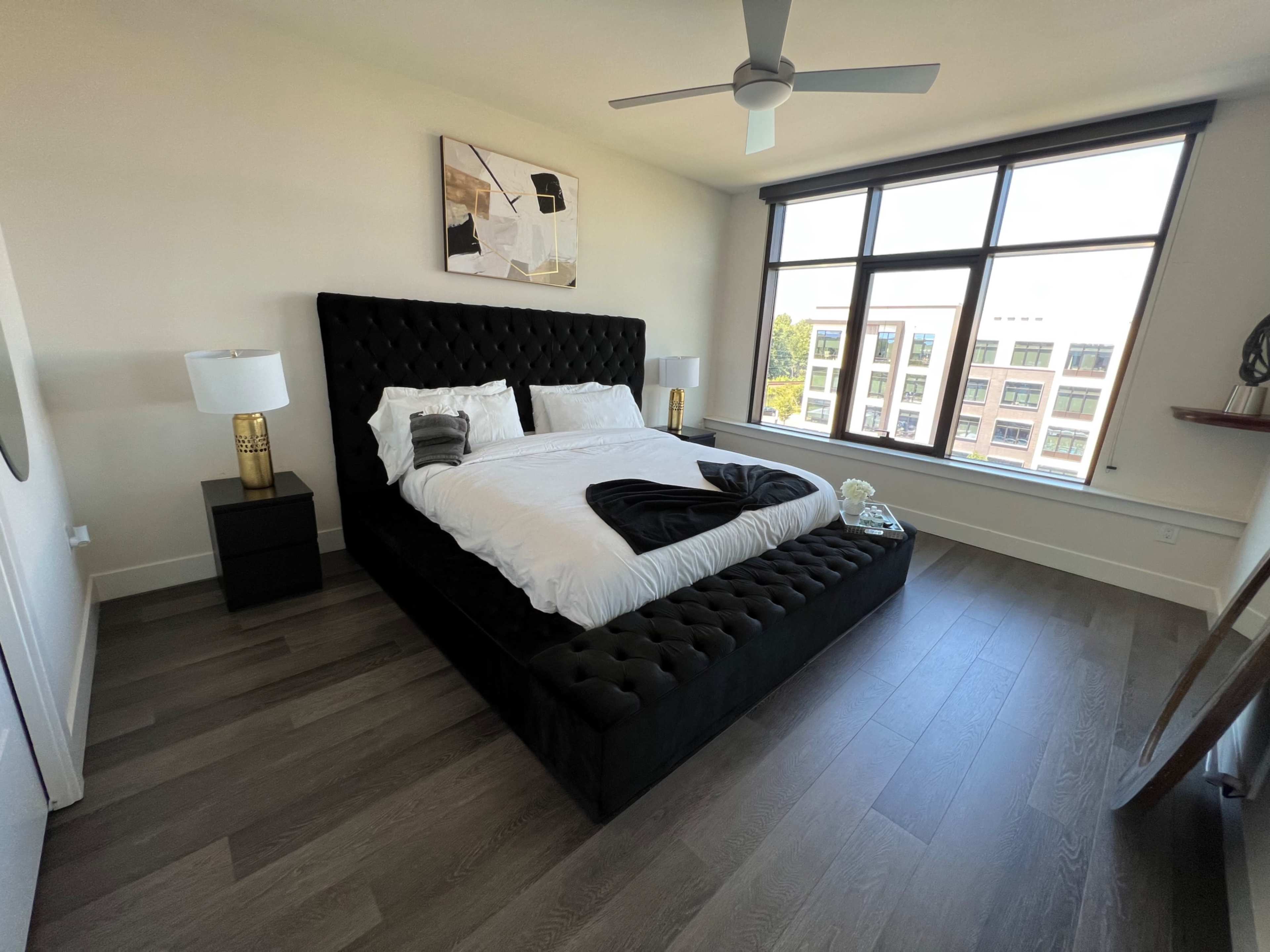 A modern bedroom featuring a large tufted black bed, two bedside lamps, and a window with a view of neighboring buildings.