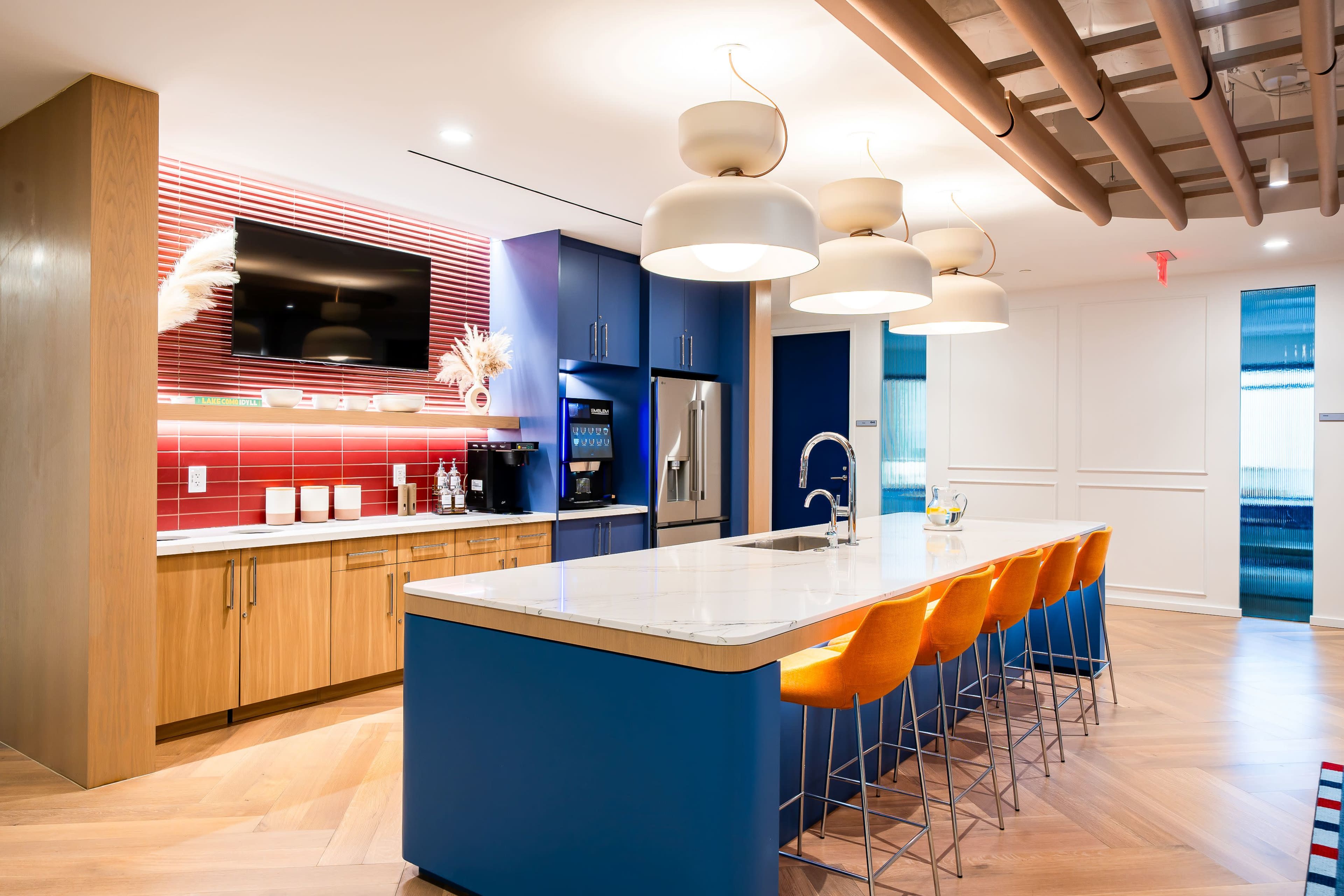 The image shows a modern kitchen with a large marble island, blue cabinetry, orange bar stools, and a wall-mounted television.