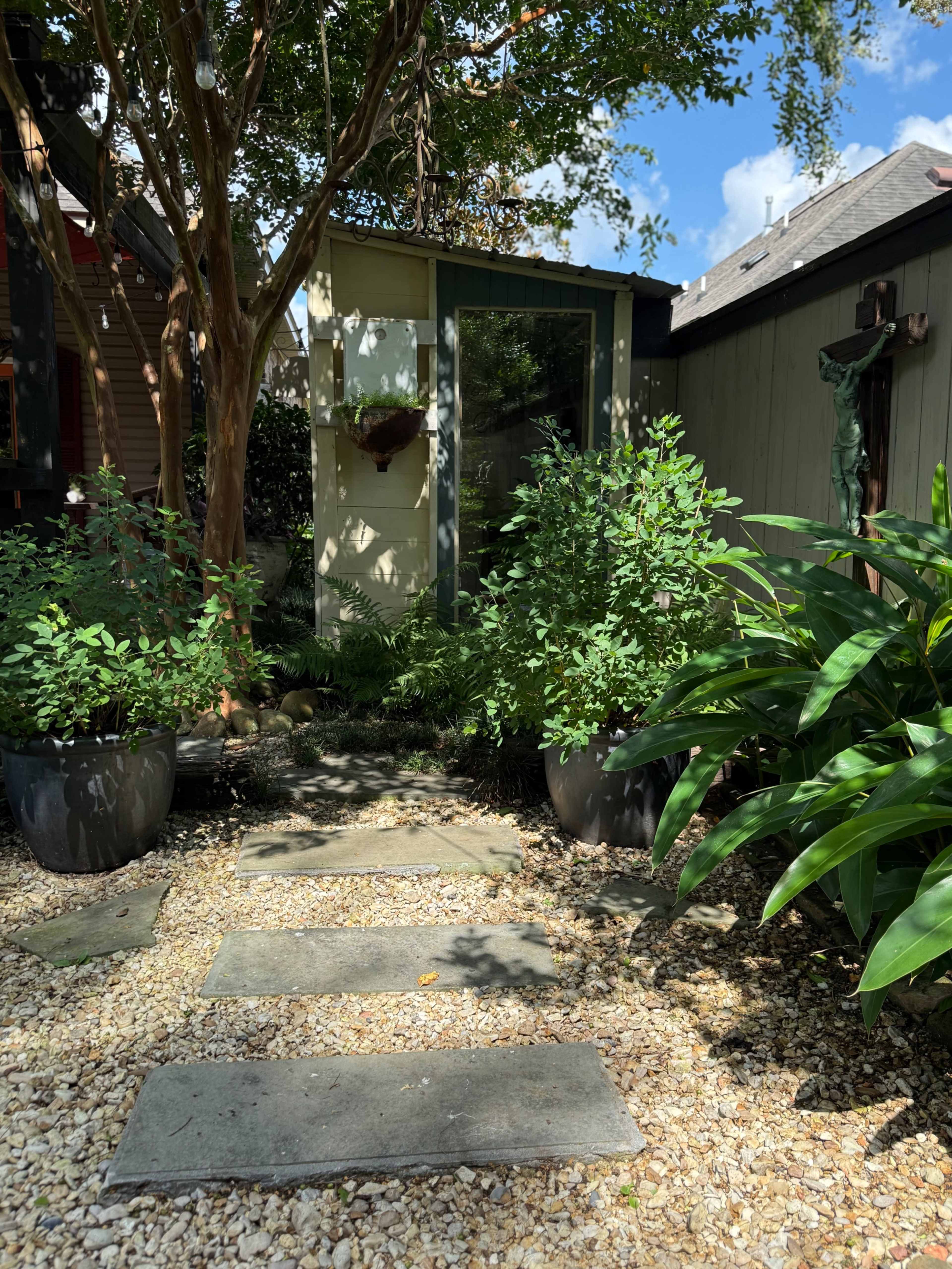 The image features a pathway of stone slabs leading to a small building, surrounded by lush green plants and decorative foliage under a clear blue sky.
