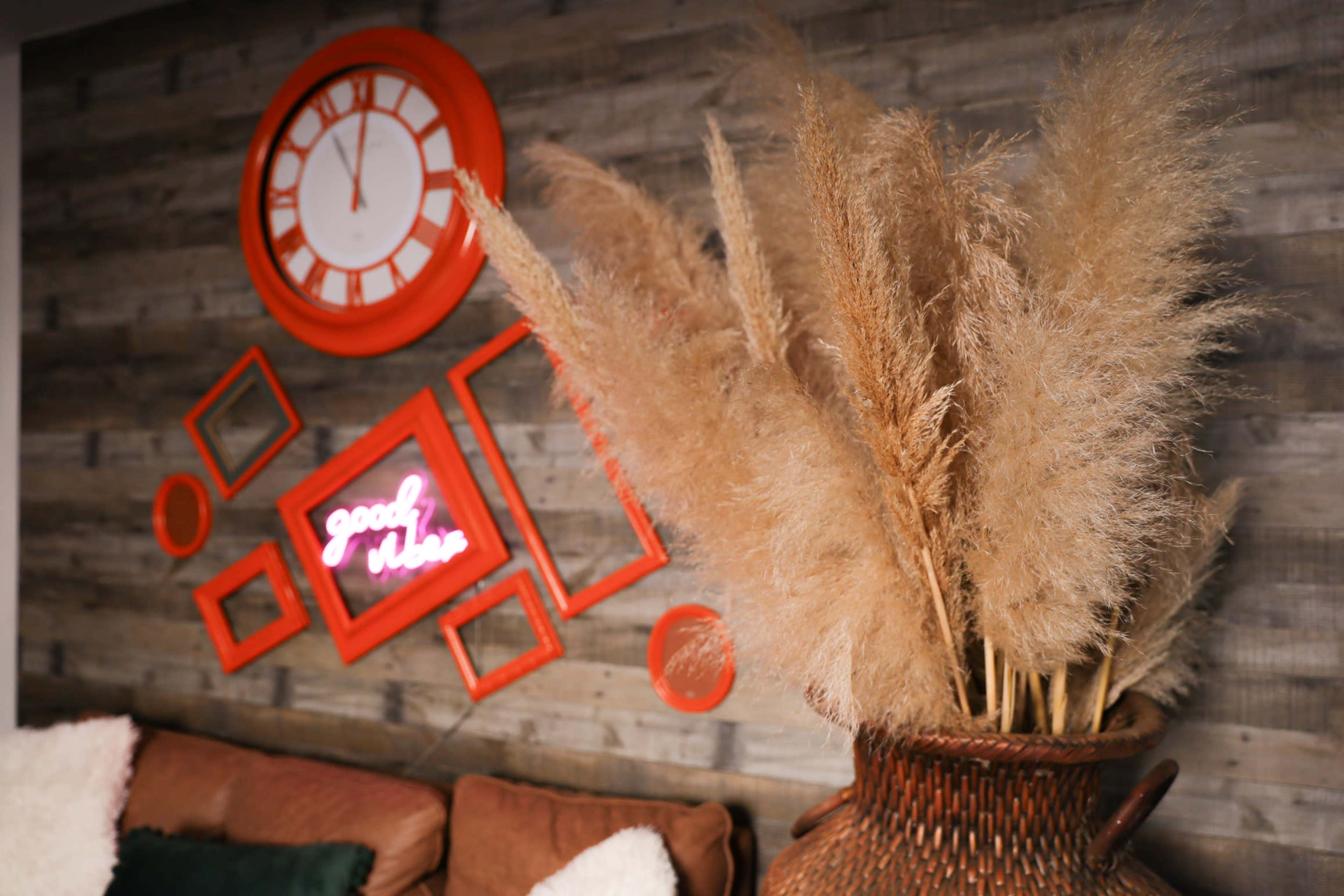 A large bouquet of dried pampas grass is displayed in a woven basket against a wooden wall featuring an orange clock and a neon sign framed in red.