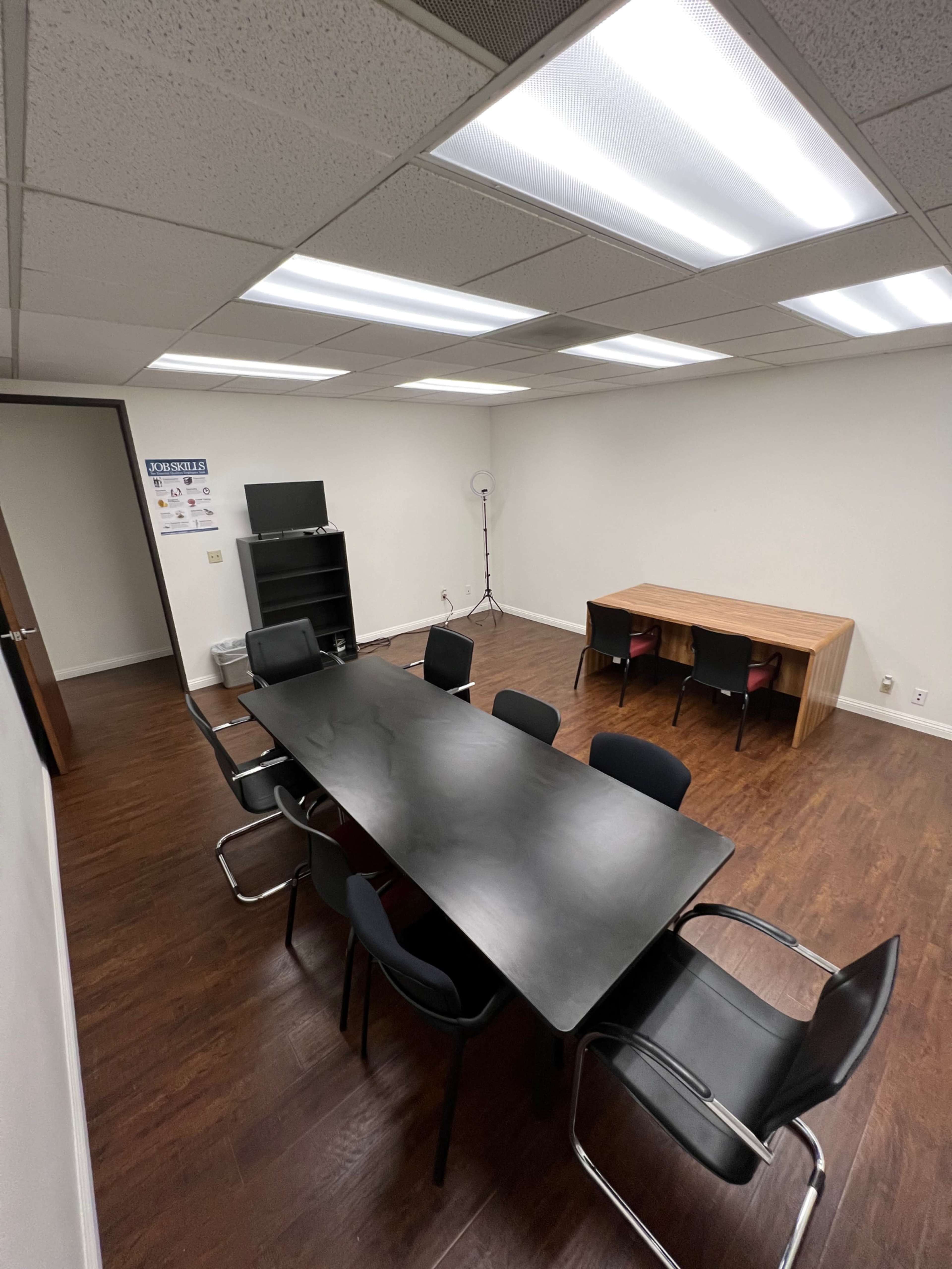 A modern office meeting room featuring a long black conference table surrounded by chairs, with a bookshelf and a wooden table in a well-lit space.