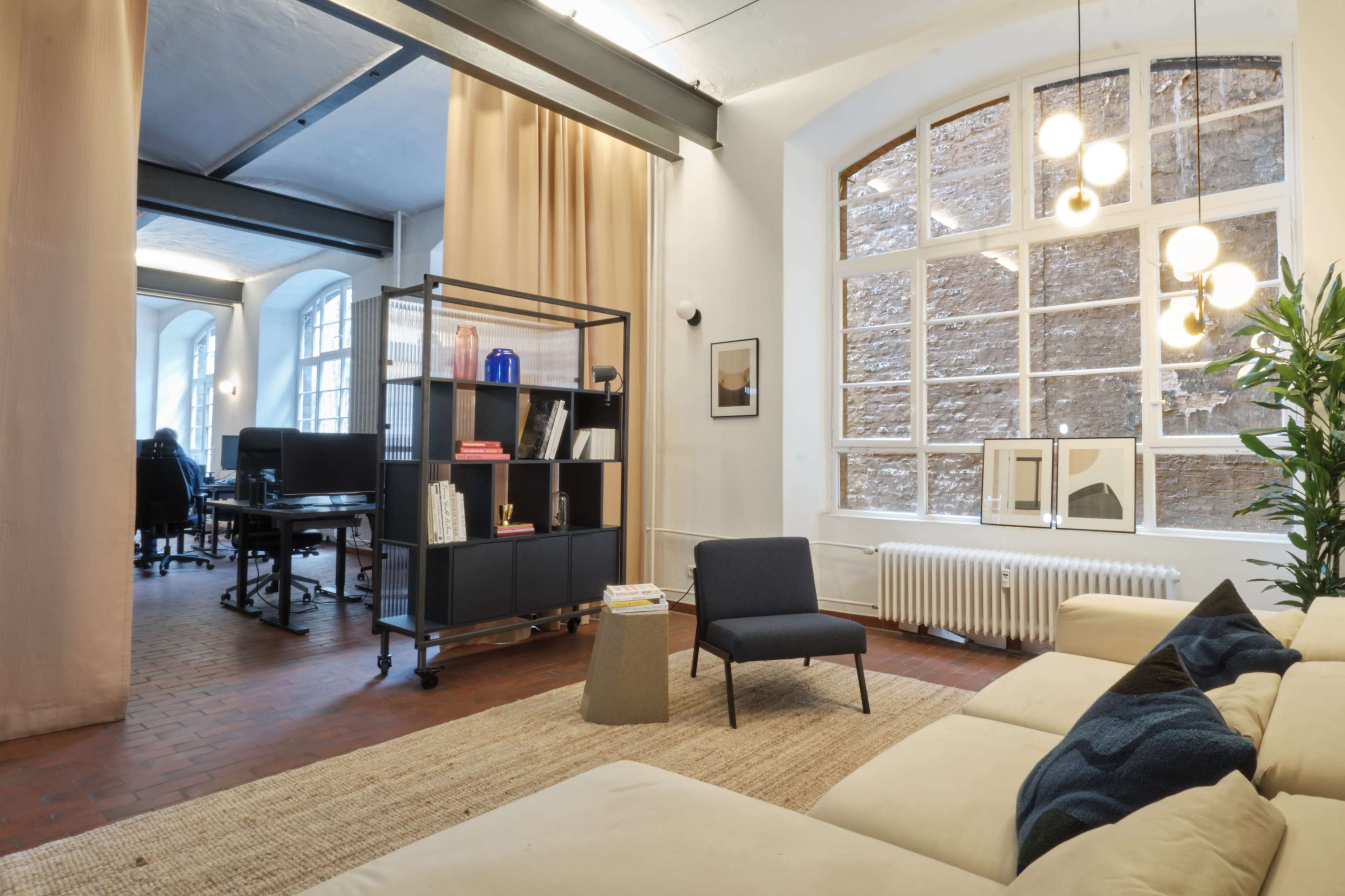 The image shows a modern open office space with a seating area, a large window with multiple panes, and a bookshelf dividing the room.