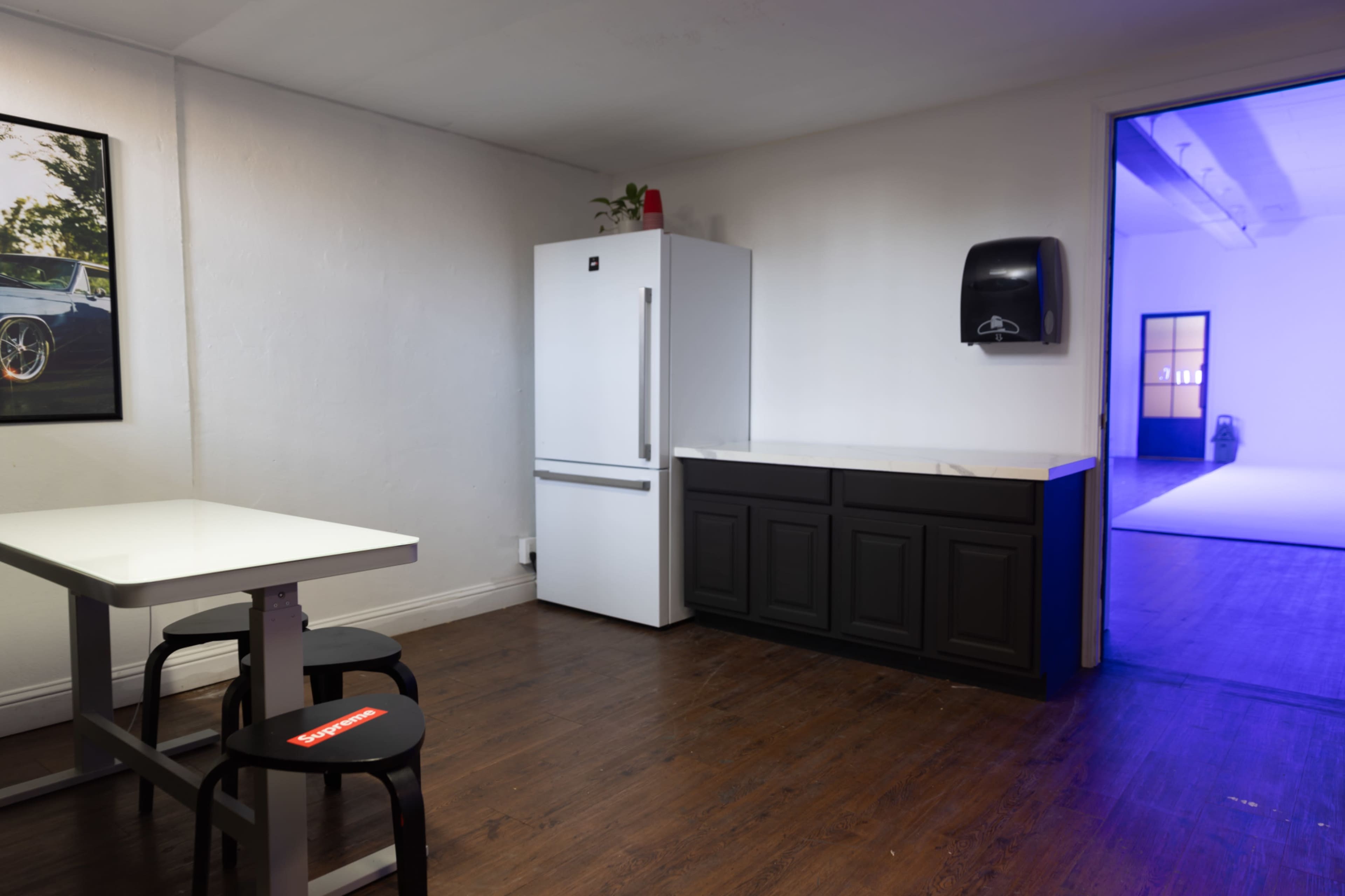 The image shows a tidy room with a white refrigerator, a dark cabinet, a table with chairs, and a doorway leading to another brightly lit space.