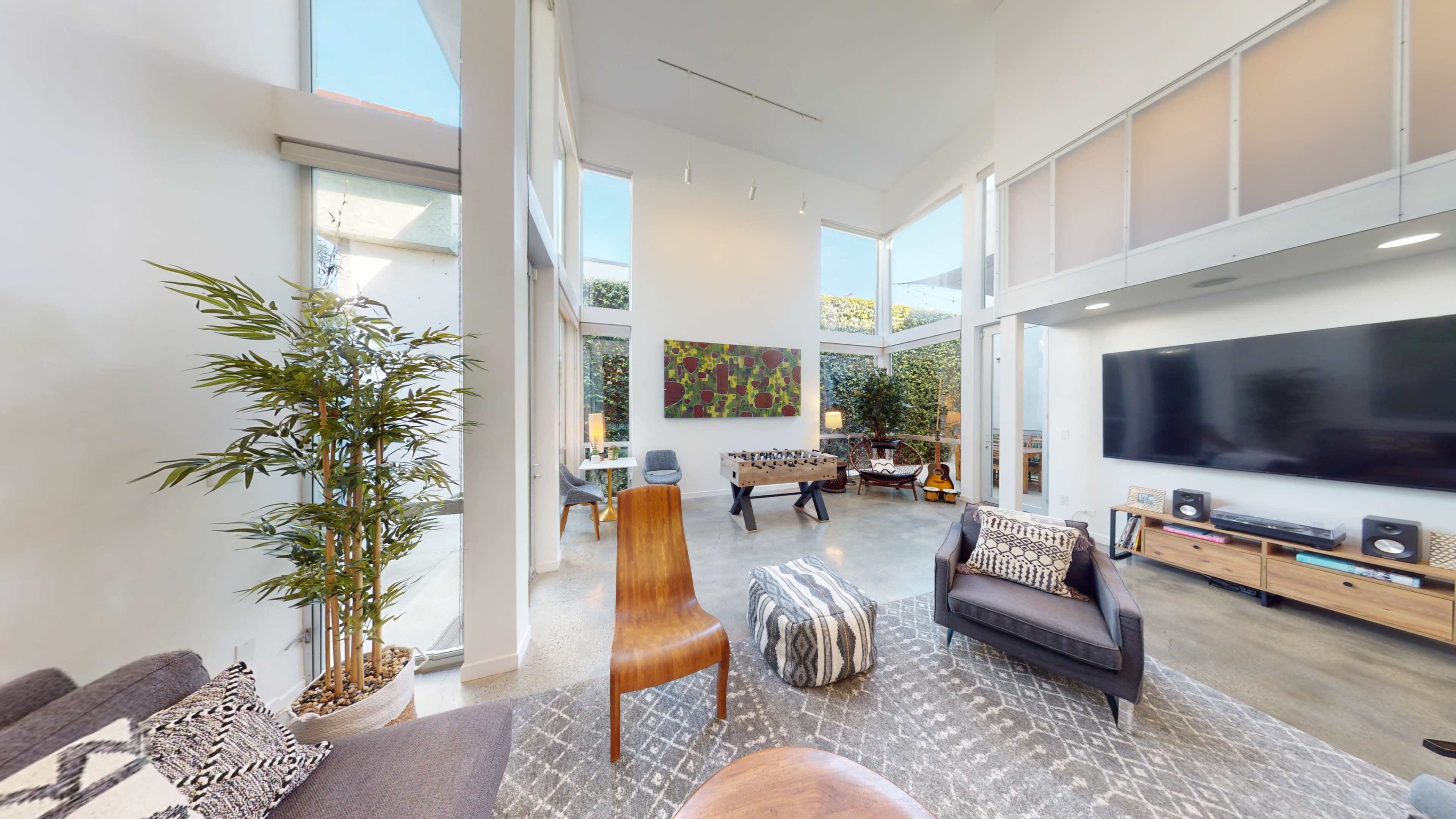 The living room features tall windows, a modern furniture arrangement, and a mixture of plants and decor elements.