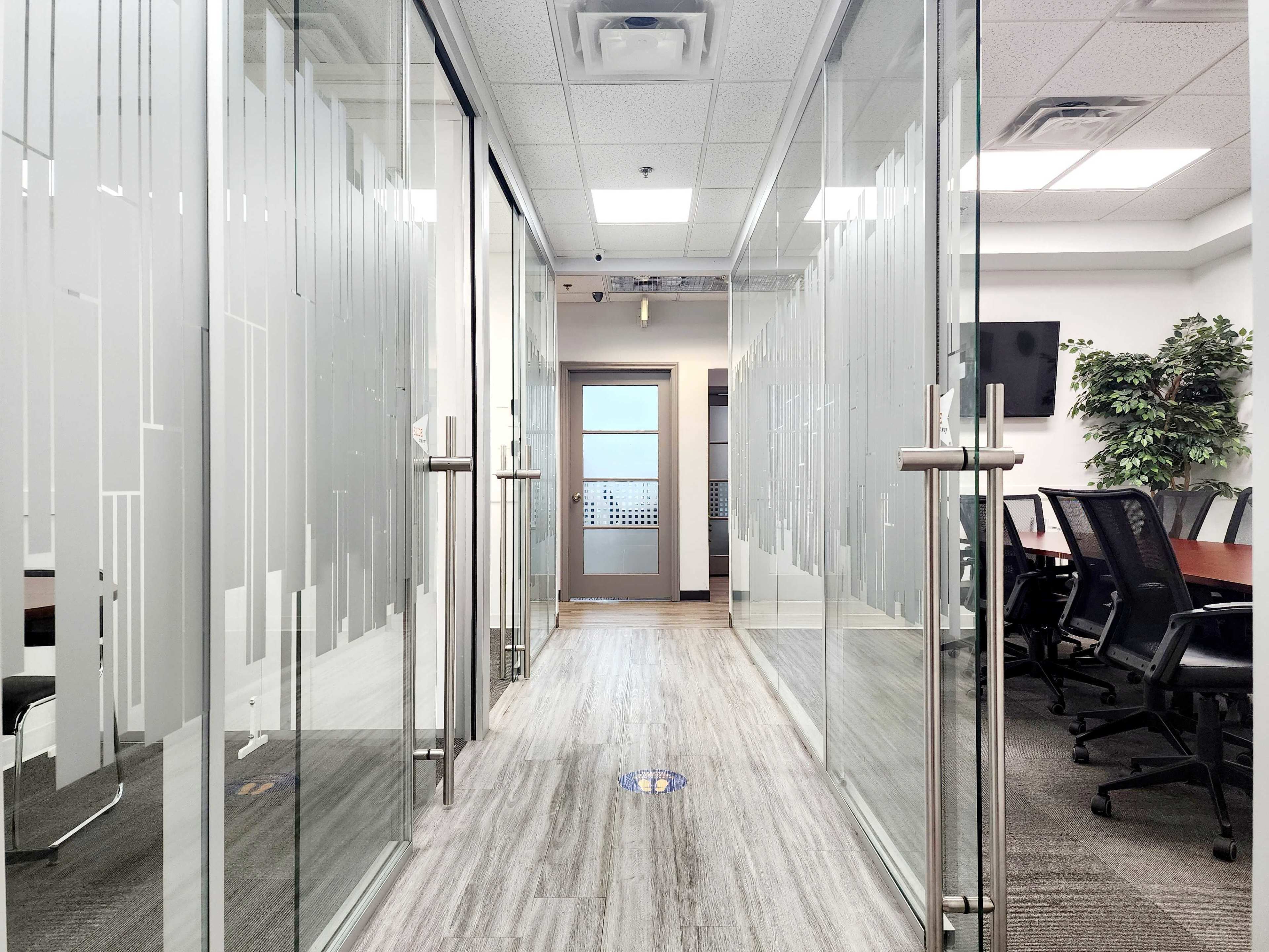 The image shows a modern office corridor with glass walls leading to a meeting room at the end.