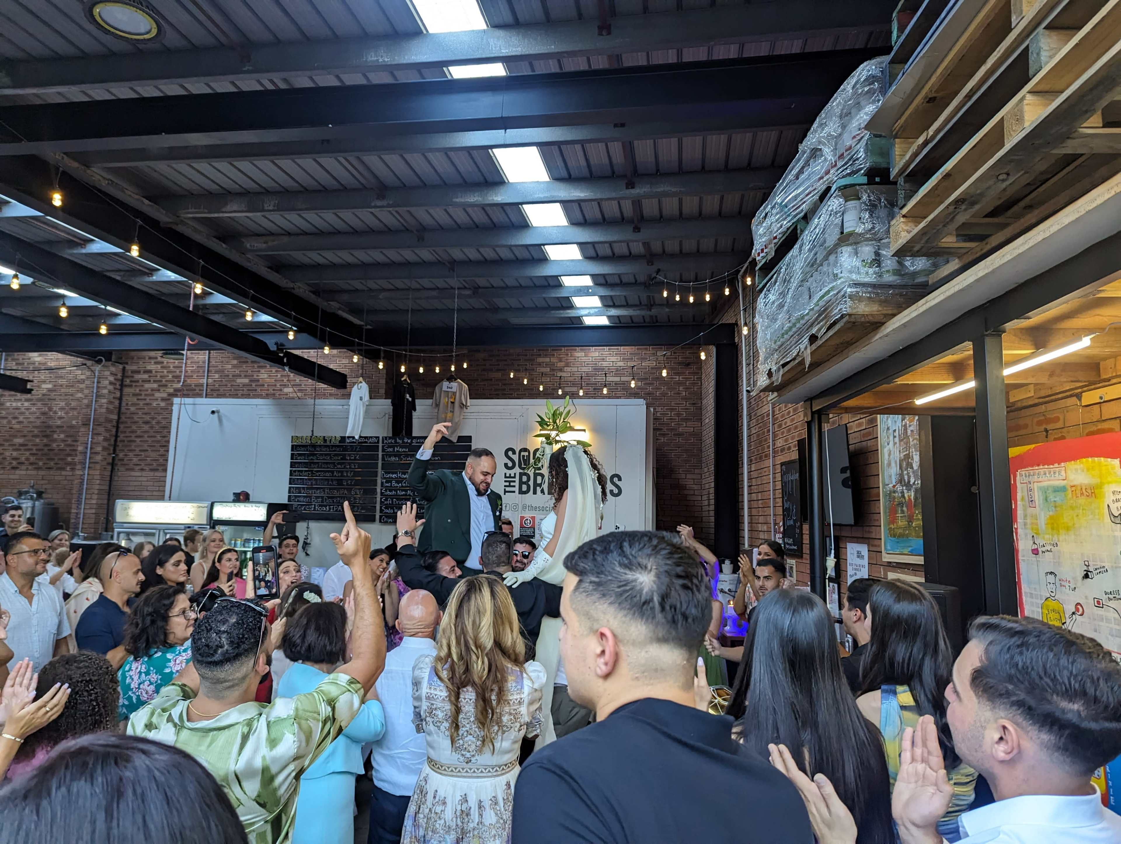A man is being lifted by guests during a celebration inside a well-lit venue with brick walls and industrial décor.
