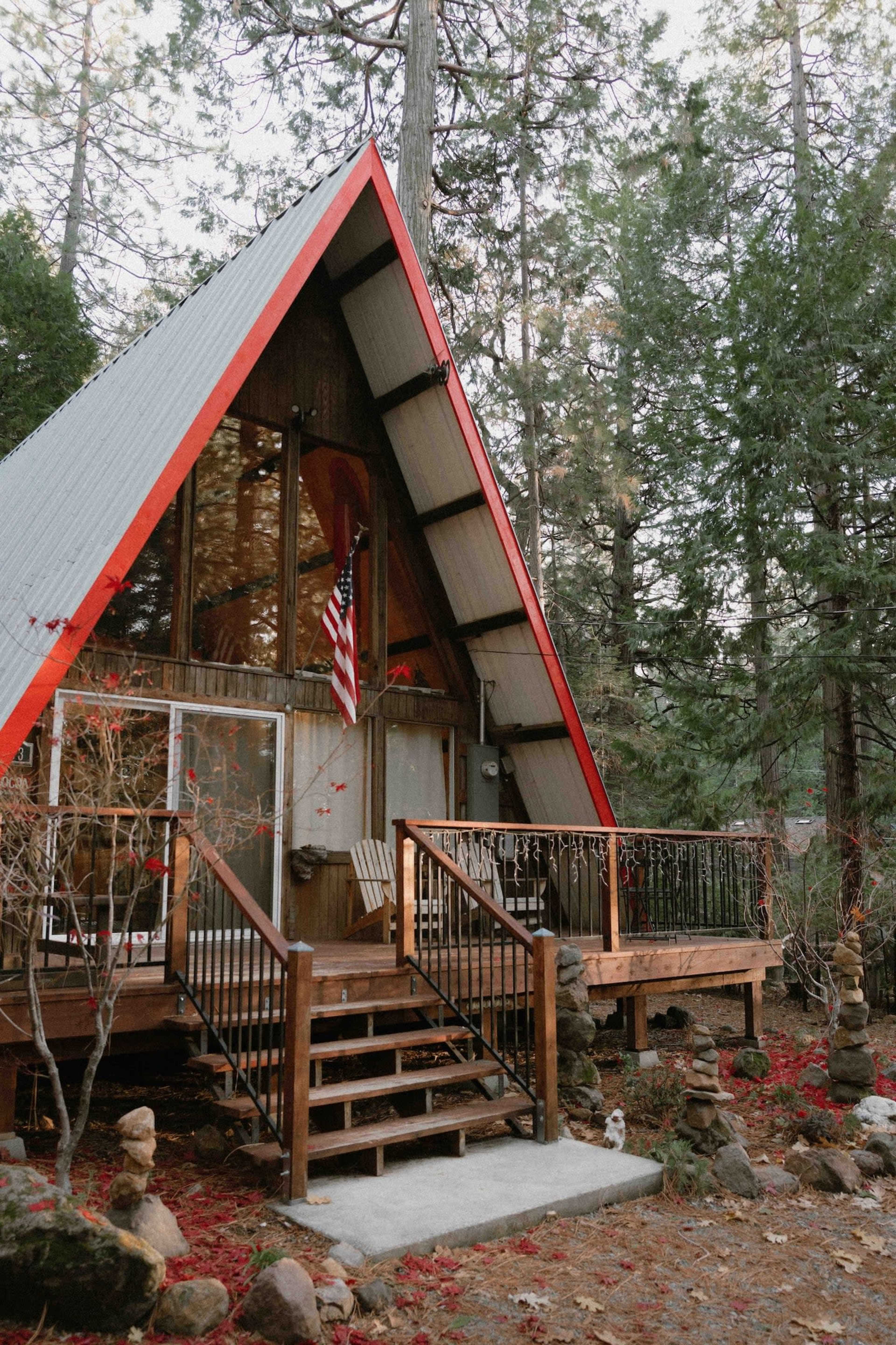An A-frame cabin with a red roof, nestled among tall trees and featuring a wooden deck with steps and an American flag.