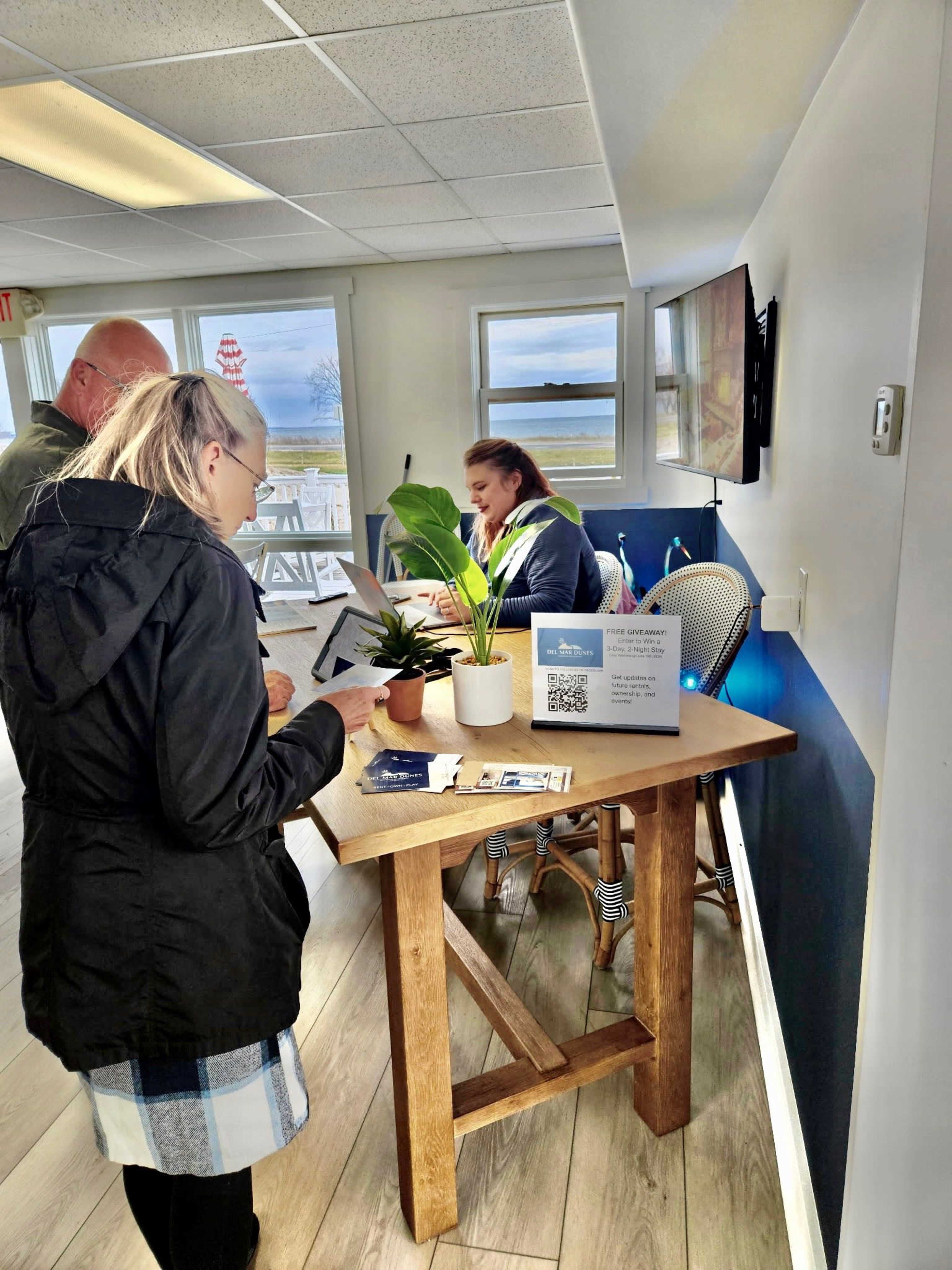 A woman is seated at a wooden table, assisting two people who are looking at brochures in a bright, modern reception area with large windows.