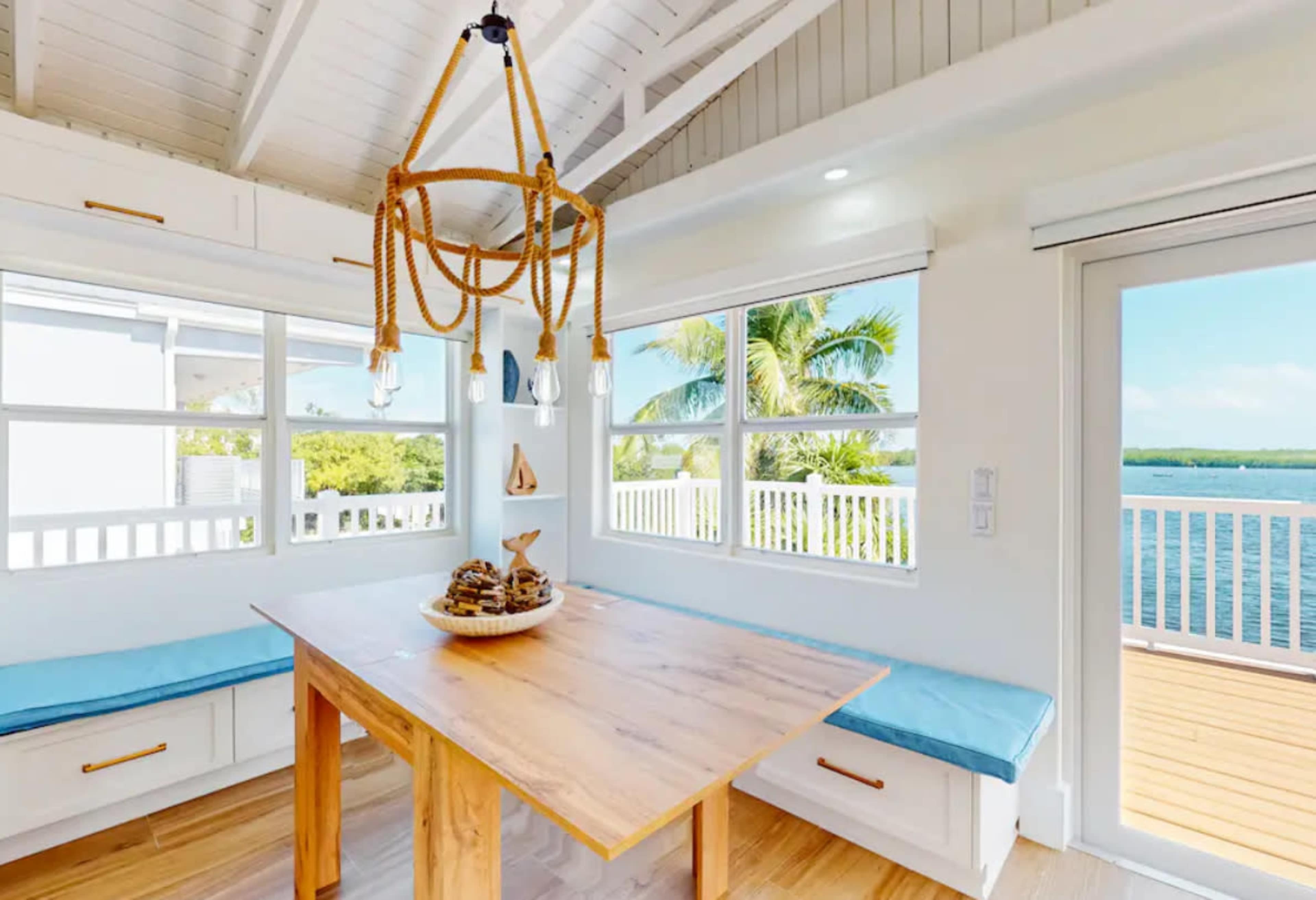 A dining area features a wooden table with a centerpiece of shells and a rope chandelier, surrounded by windows offering a view of the water.