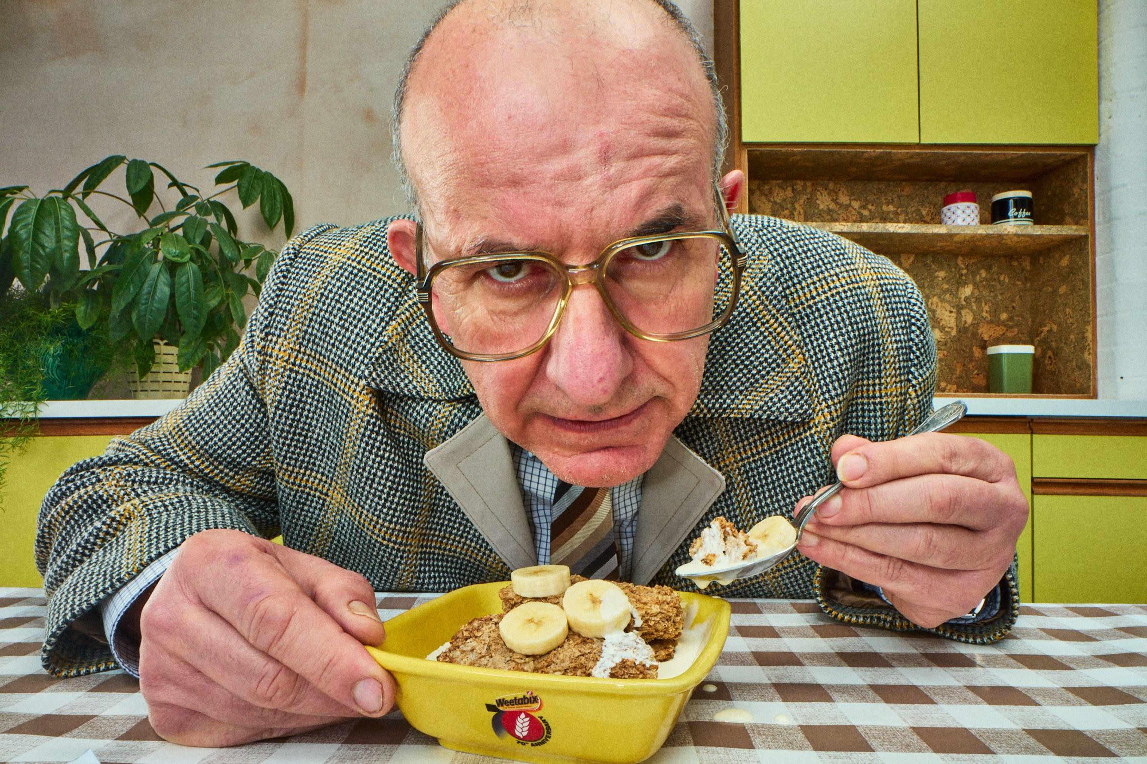 A man in a plaid suit sits at a checkered table, intently eating a bowl of cereal topped with banana slices.