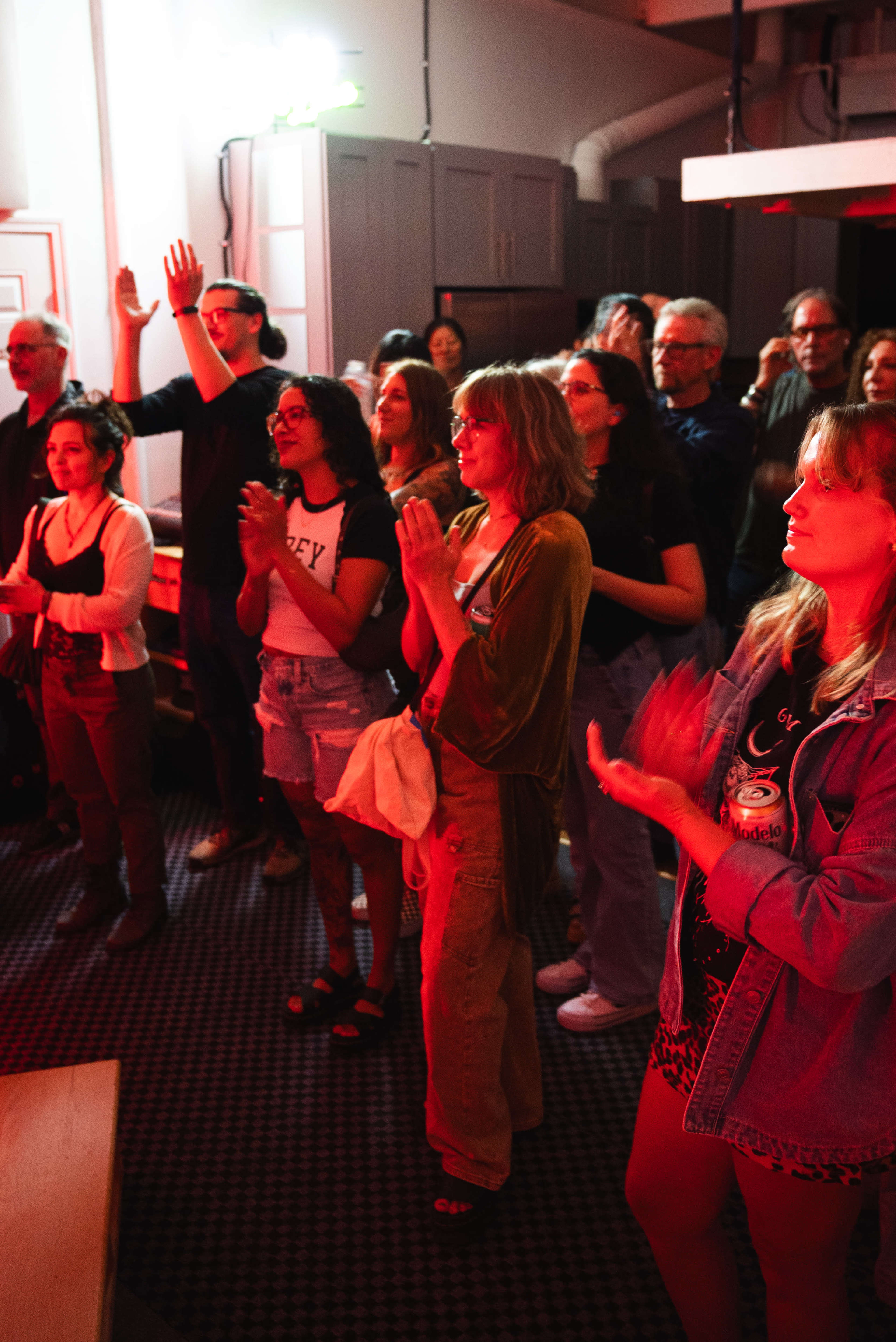 A diverse group of people is standing and clapping in a crowded indoor venue with dim lighting.