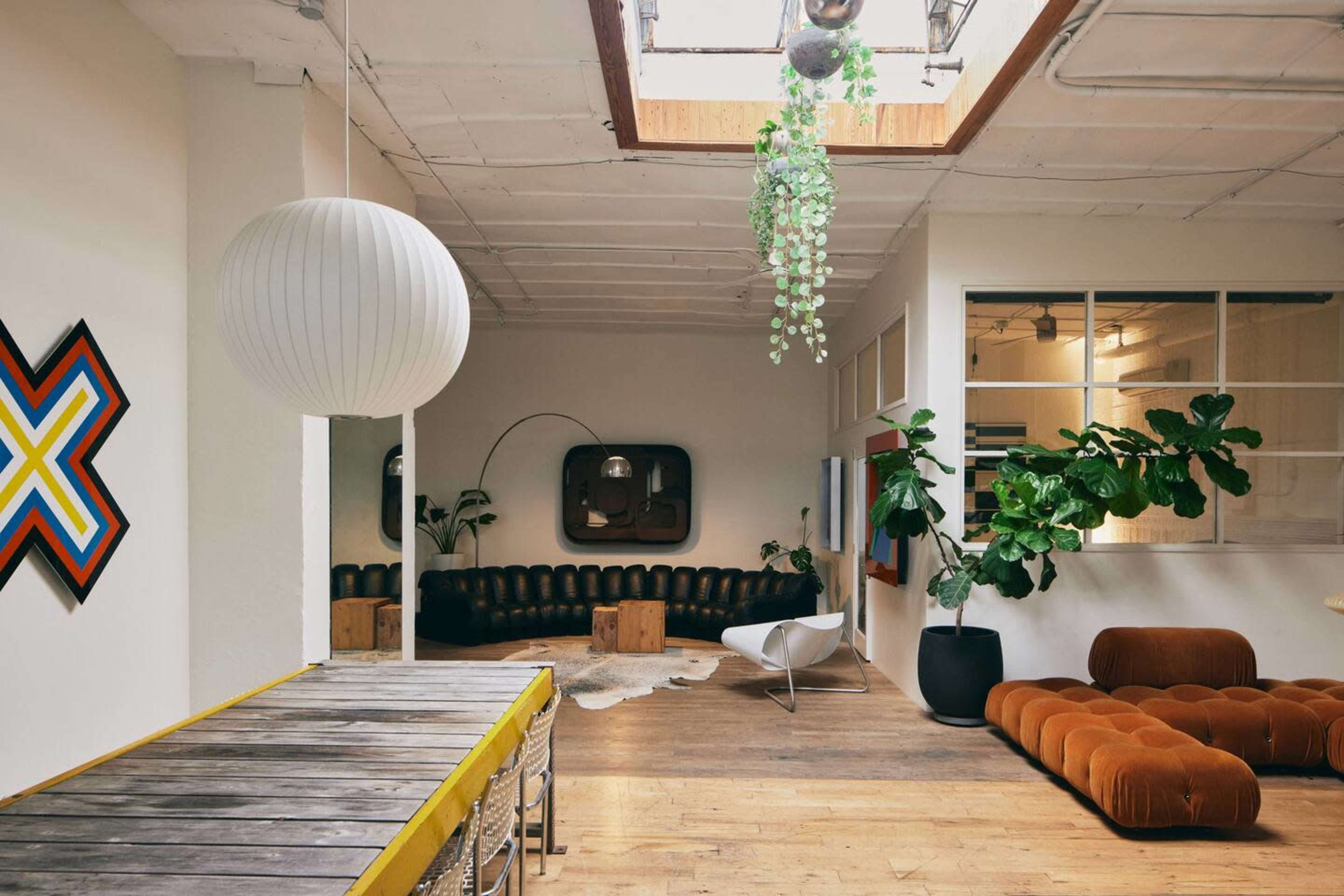 The image shows a spacious, well-lit interior with a mix of modern furniture, plants, and a skylight illuminating the room.