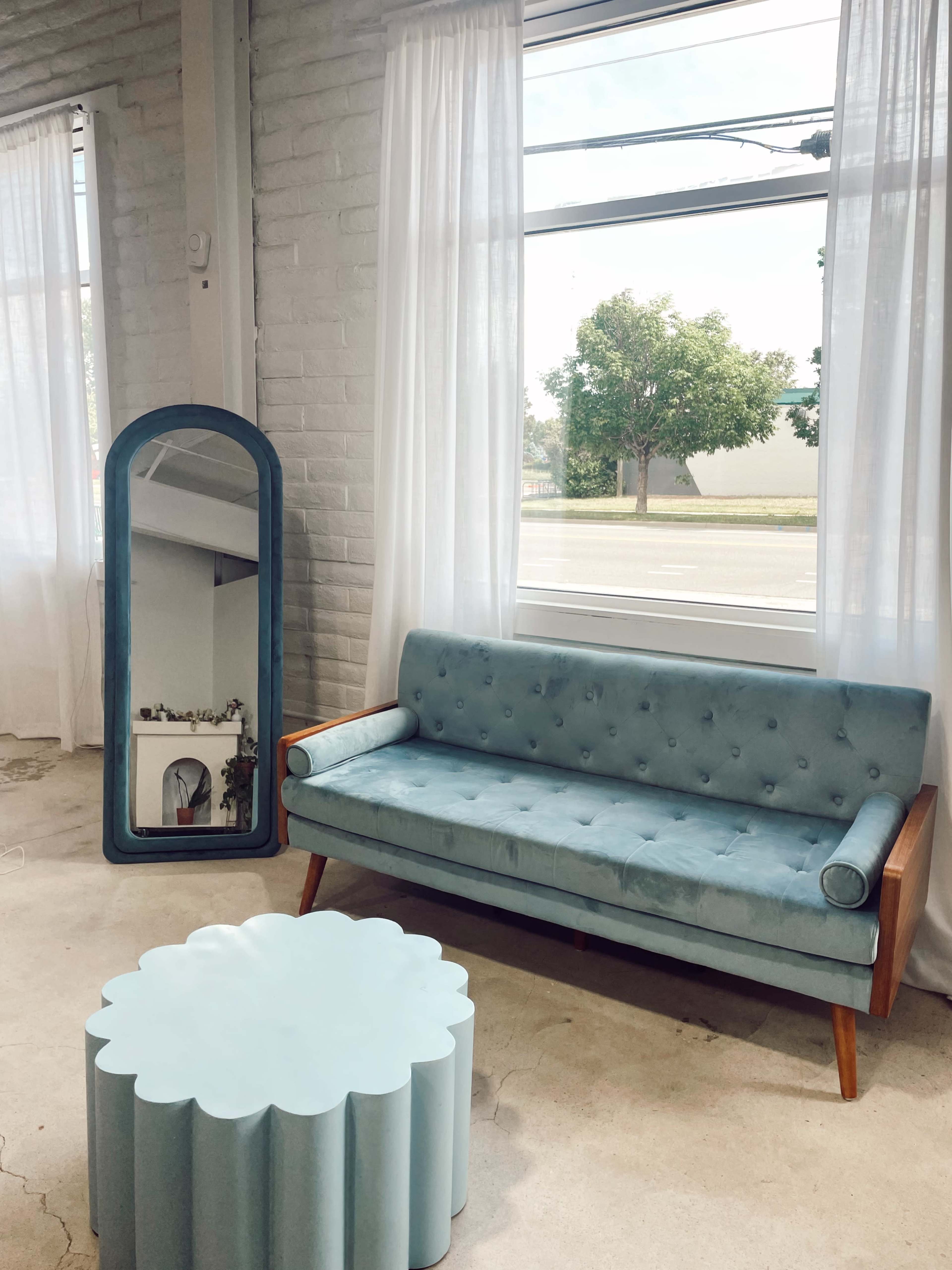 A turquoise couch with wooden legs is positioned next to a round, scalloped coffee table in a bright room with large windows and sheer white curtains.
