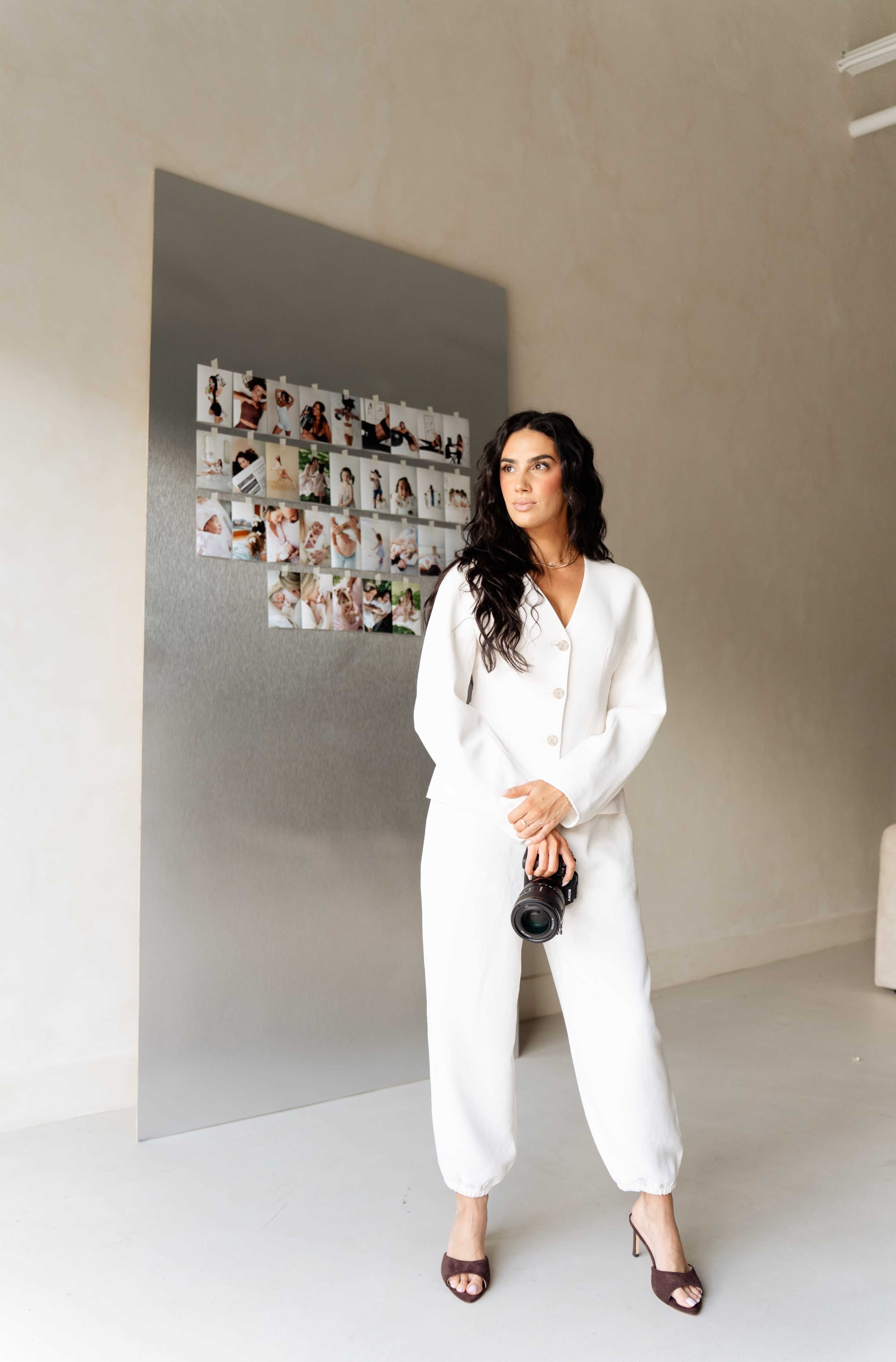 A woman in a white outfit holds a camera and stands in an indoor space with a photo collage displayed on a wall behind her.
