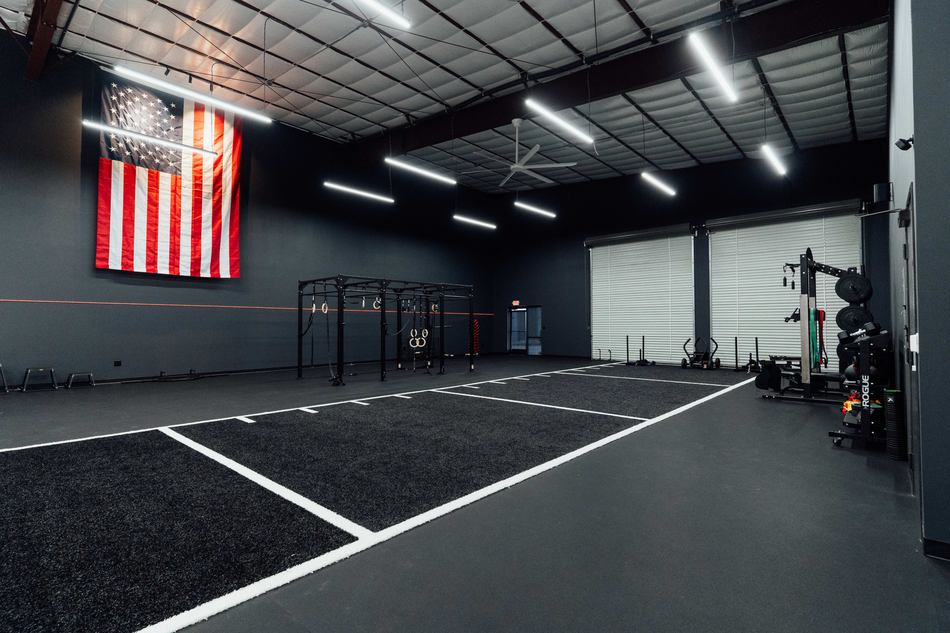 An indoor gym featuring turf flooring, marked exercise areas, a black training rig, and an American flag displayed on the wall.