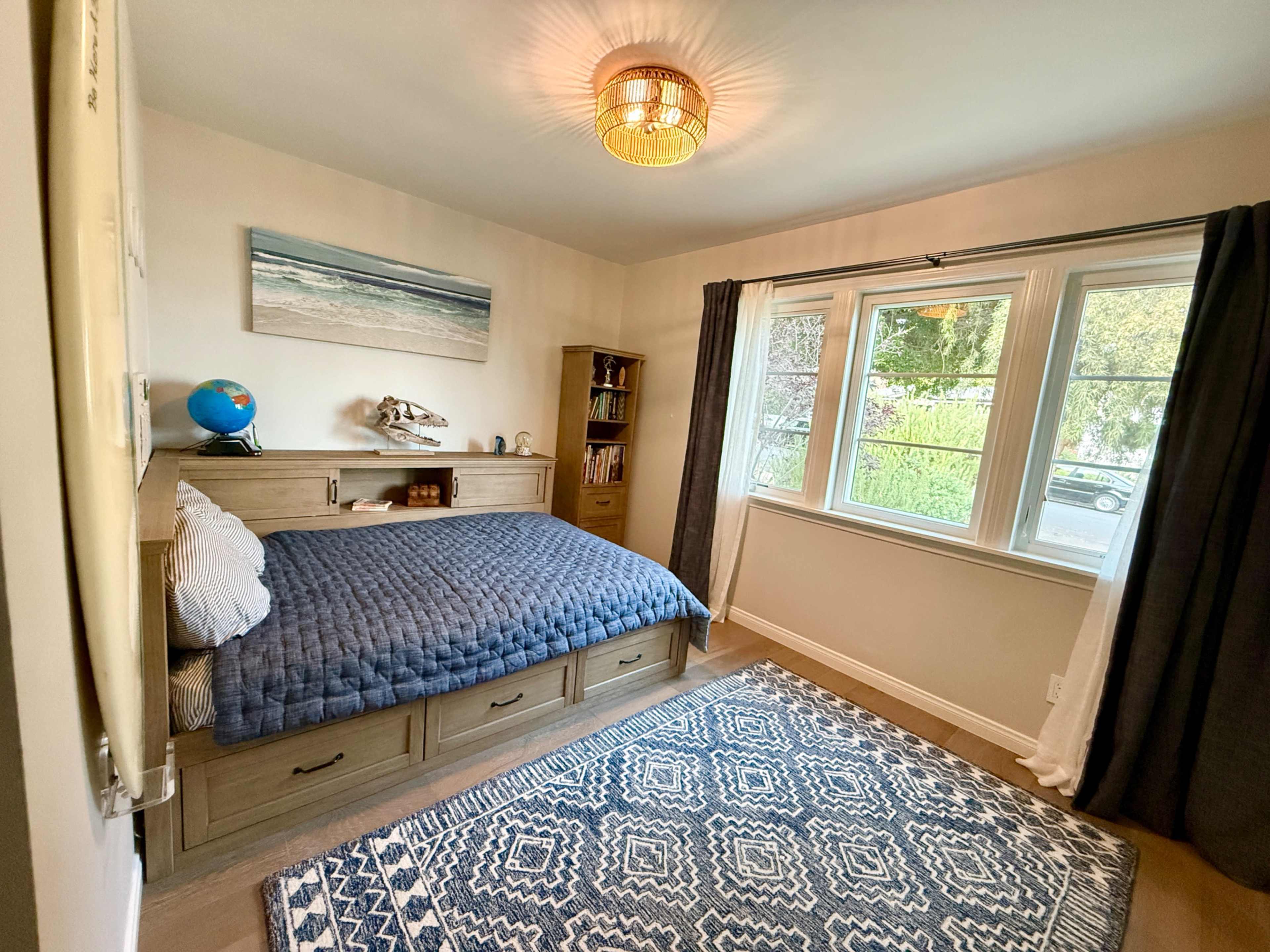 The image shows a cozy bedroom featuring a bed with storage drawers, a wooden bookshelf, a globe, and a window with curtains, allowing natural light to illuminate the space.