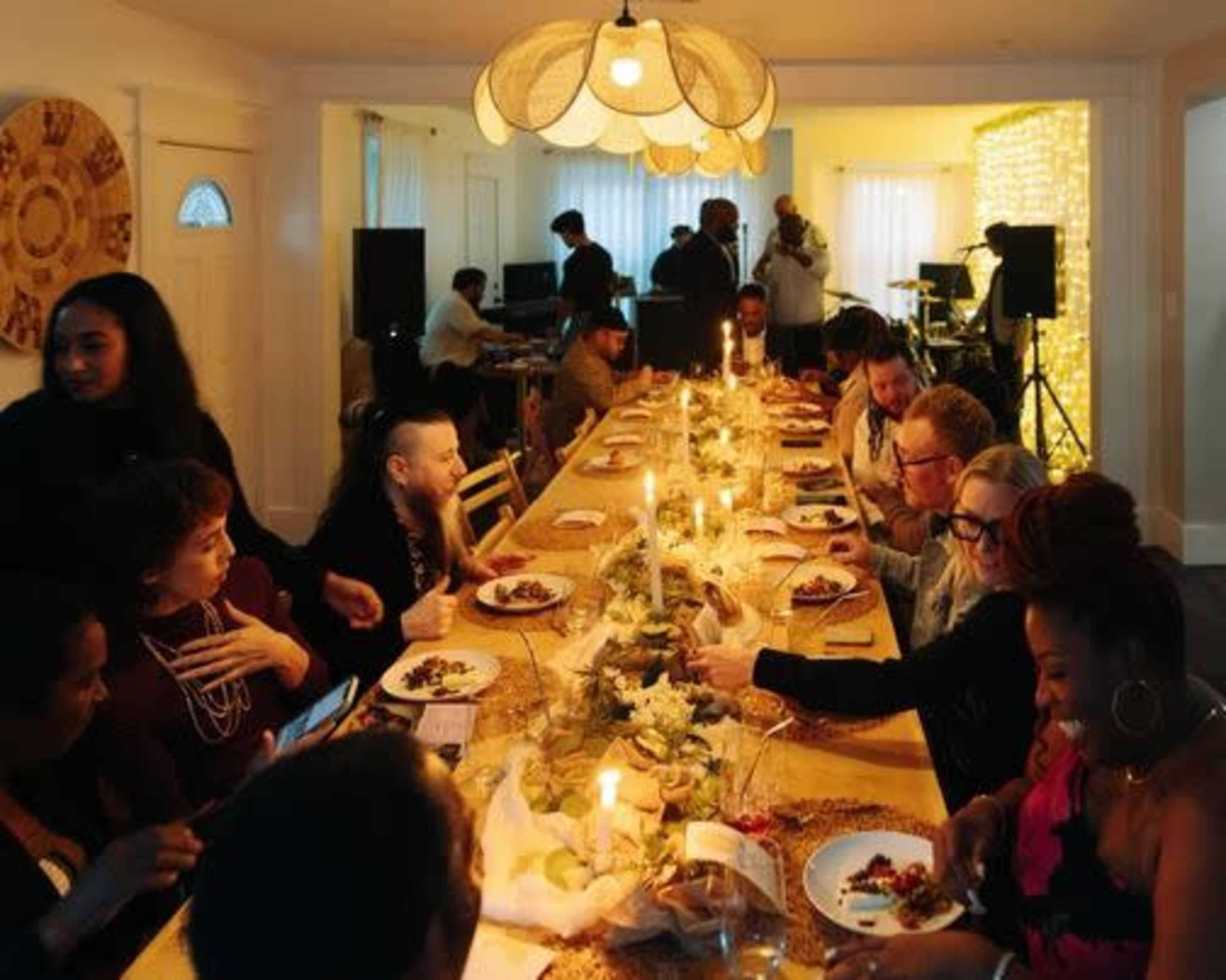 A long dining table is set with plates of food and candles, while a group of people enjoy a meal and converse in a brightly lit room.