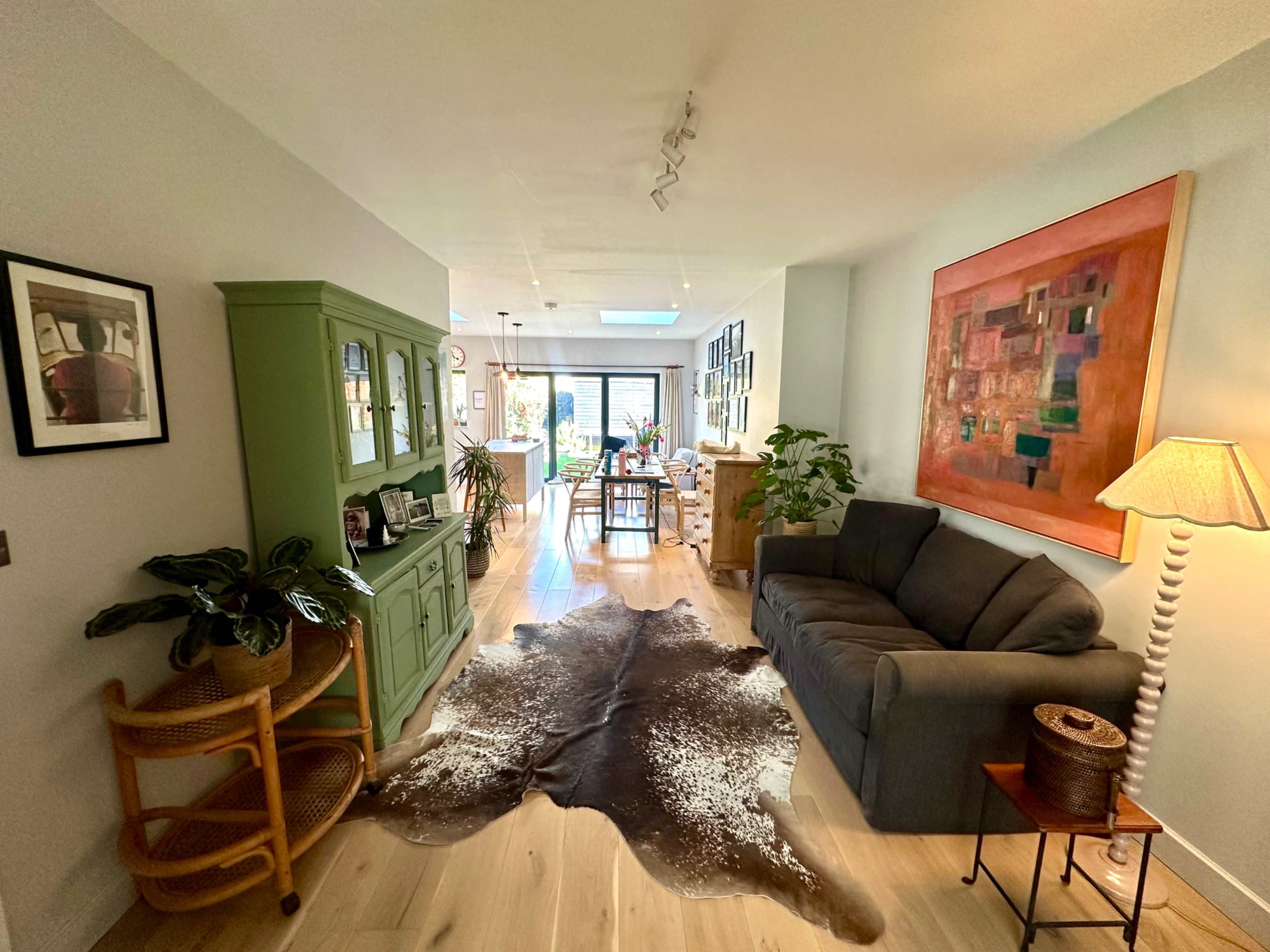 A brightly lit hallway features a green cabinet, a gray sofa, and a decorative cowhide rug, leading to a dining area in the background.