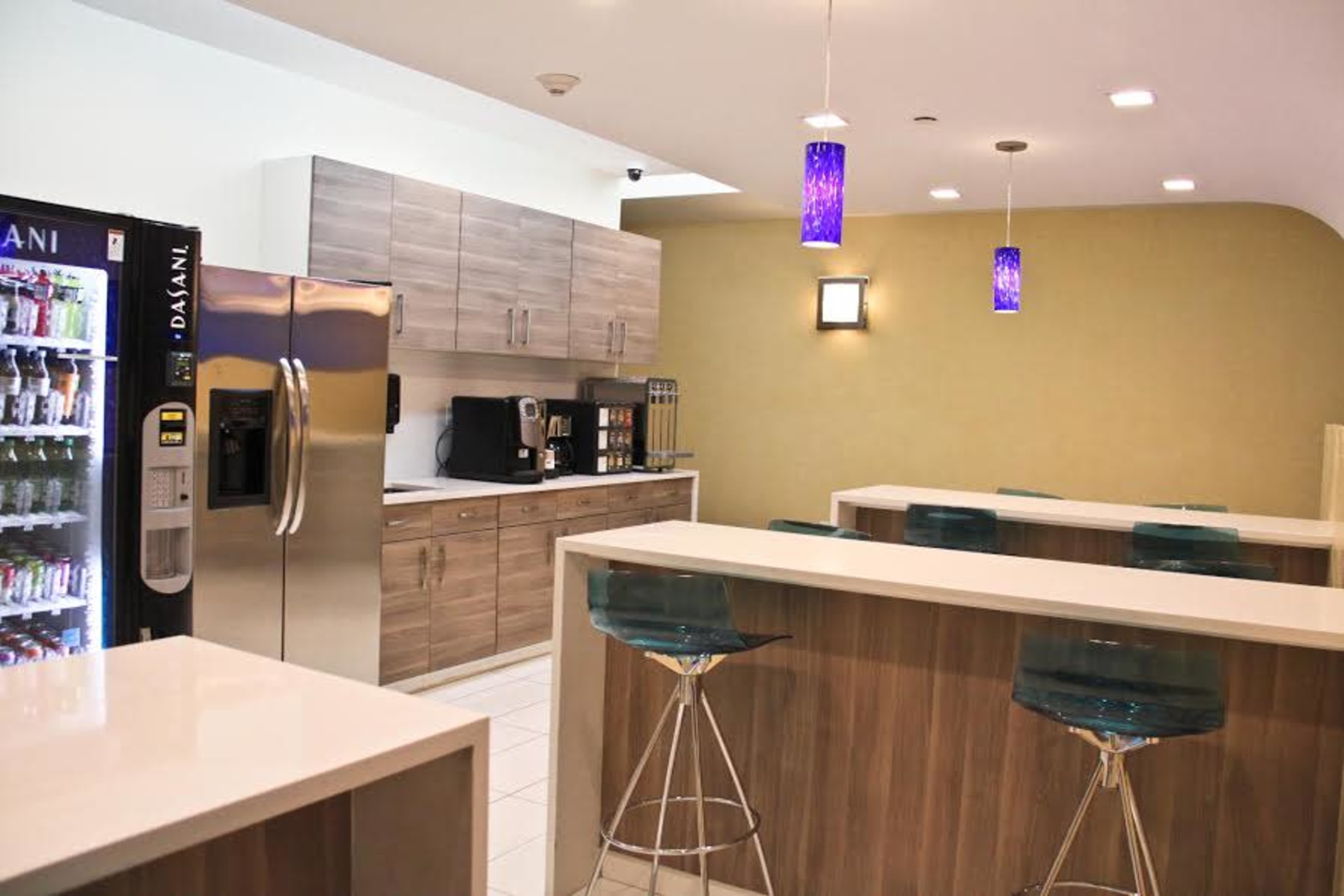The image shows a modern break room with light wooden cabinets, a stainless steel refrigerator, a coffee station, and a seating area with translucent blue bar stools.
