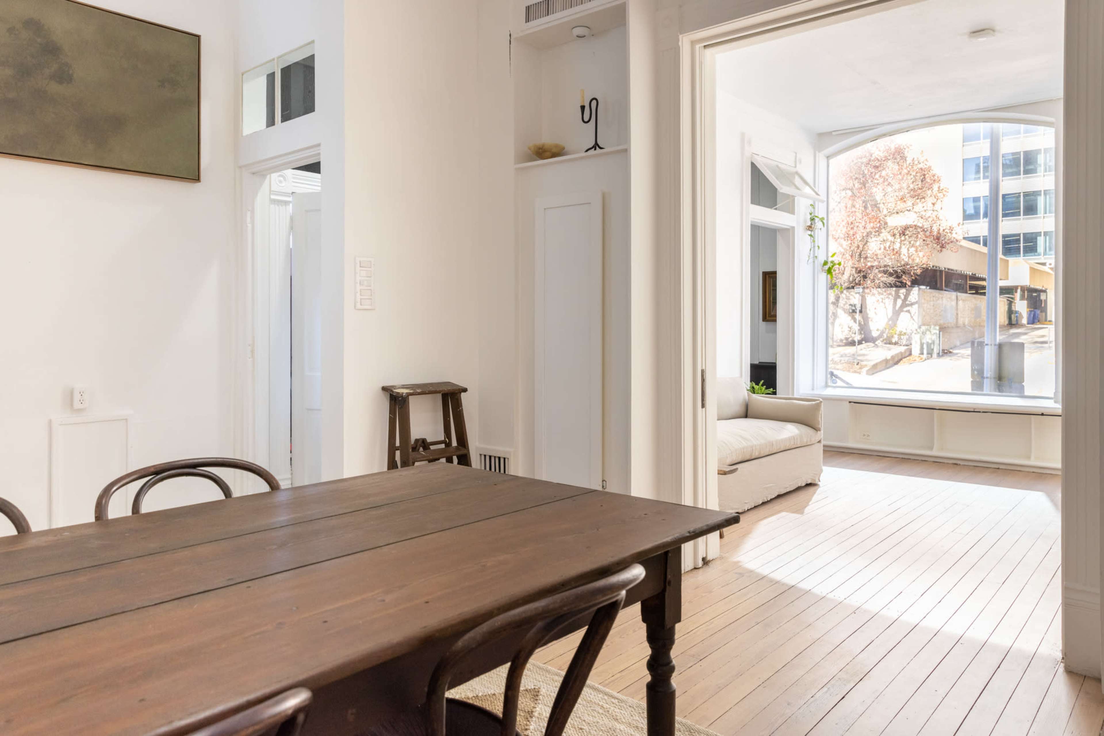 A wooden dining table in the foreground with a doorway leading to a bright living room featuring a large window.