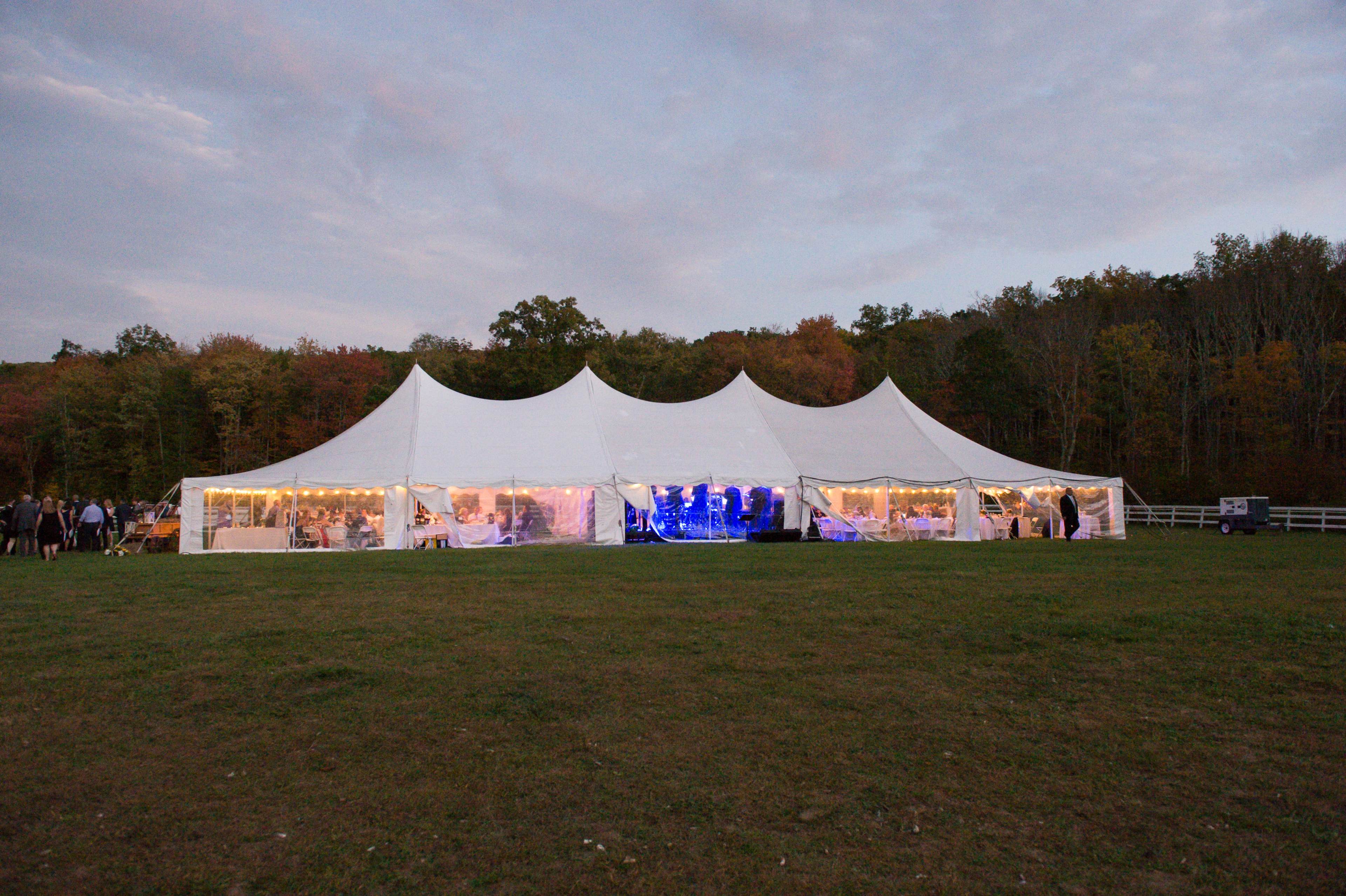 A large white tent is set up on a grassy field, with strings of lights illuminating the interior against a backdrop of trees and an evening sky.