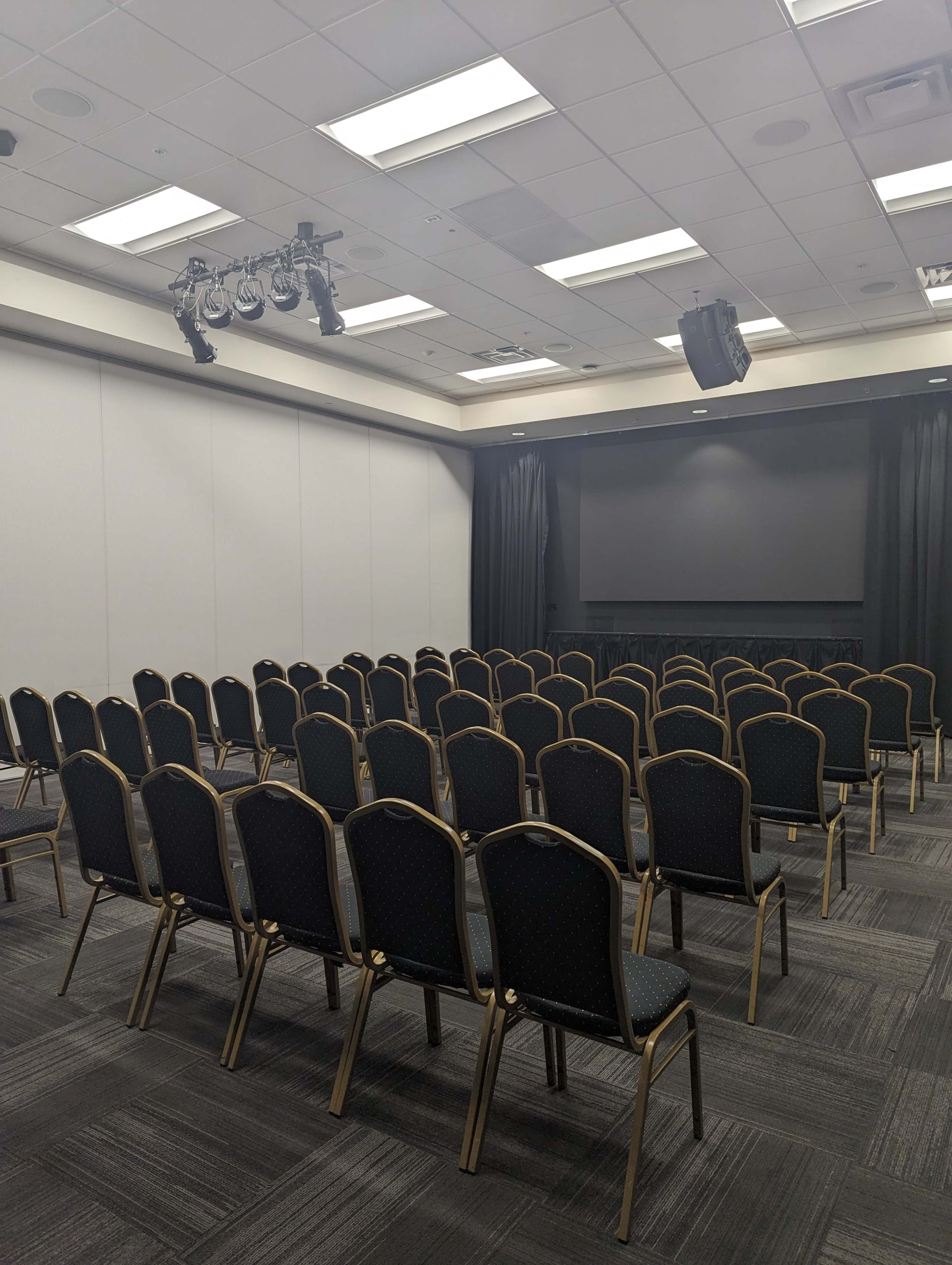 A conference room set up with rows of empty black chairs facing a stage area with curtains and lighting.