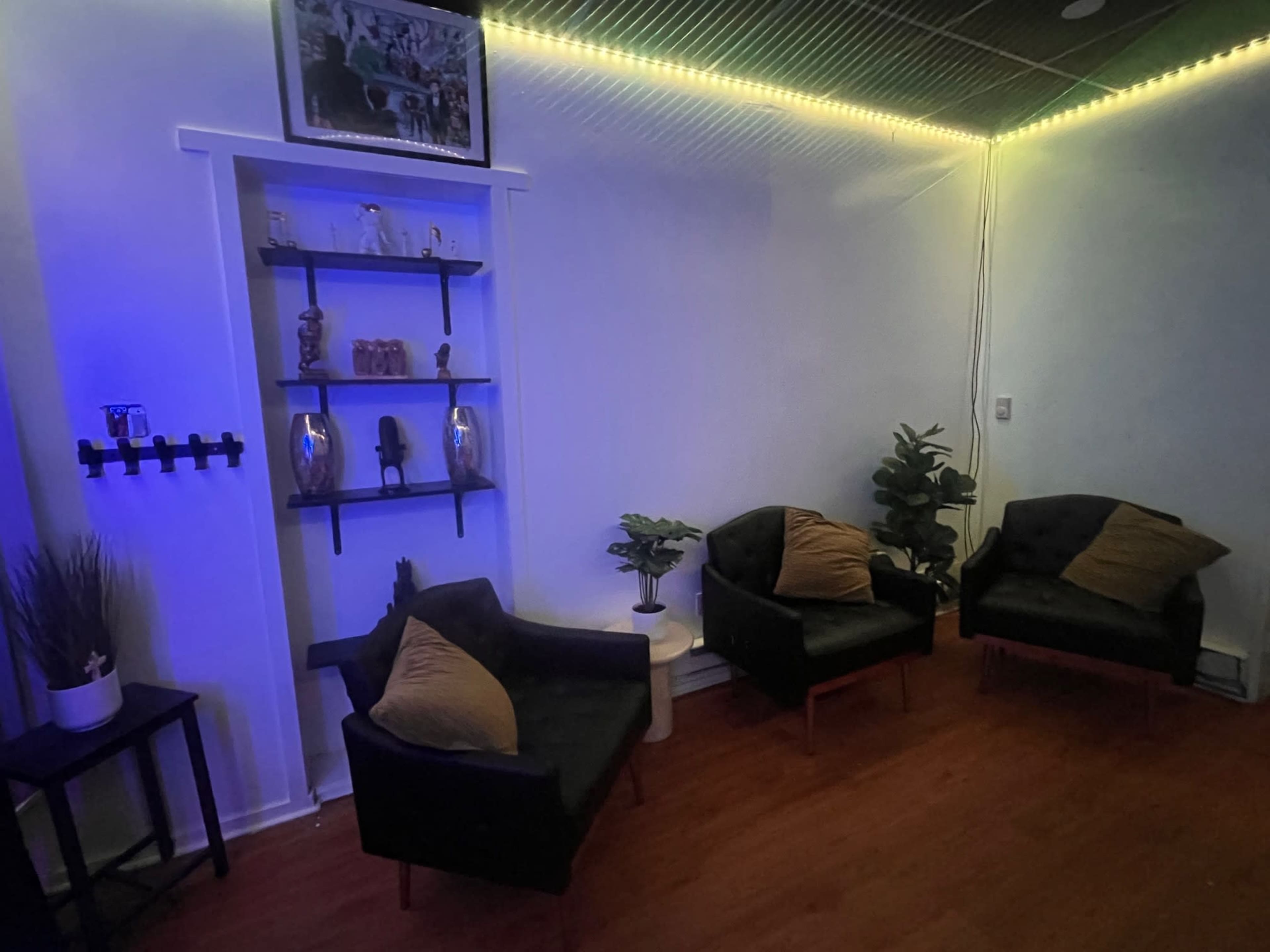 The image shows a cozy room with two black armchairs and a small table, illuminated by soft blue lighting, along with a plant and decorative shelves on the wall.