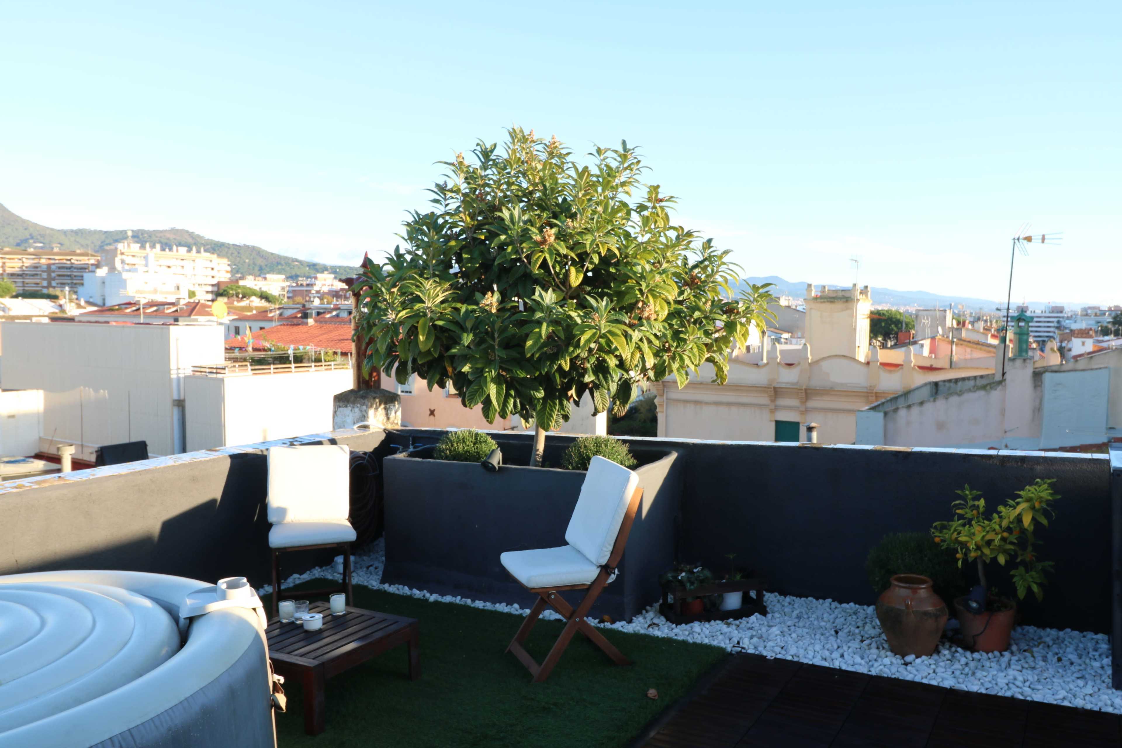 The image shows a rooftop terrace with a small seating area, a tree in a planter, and decorative stones.