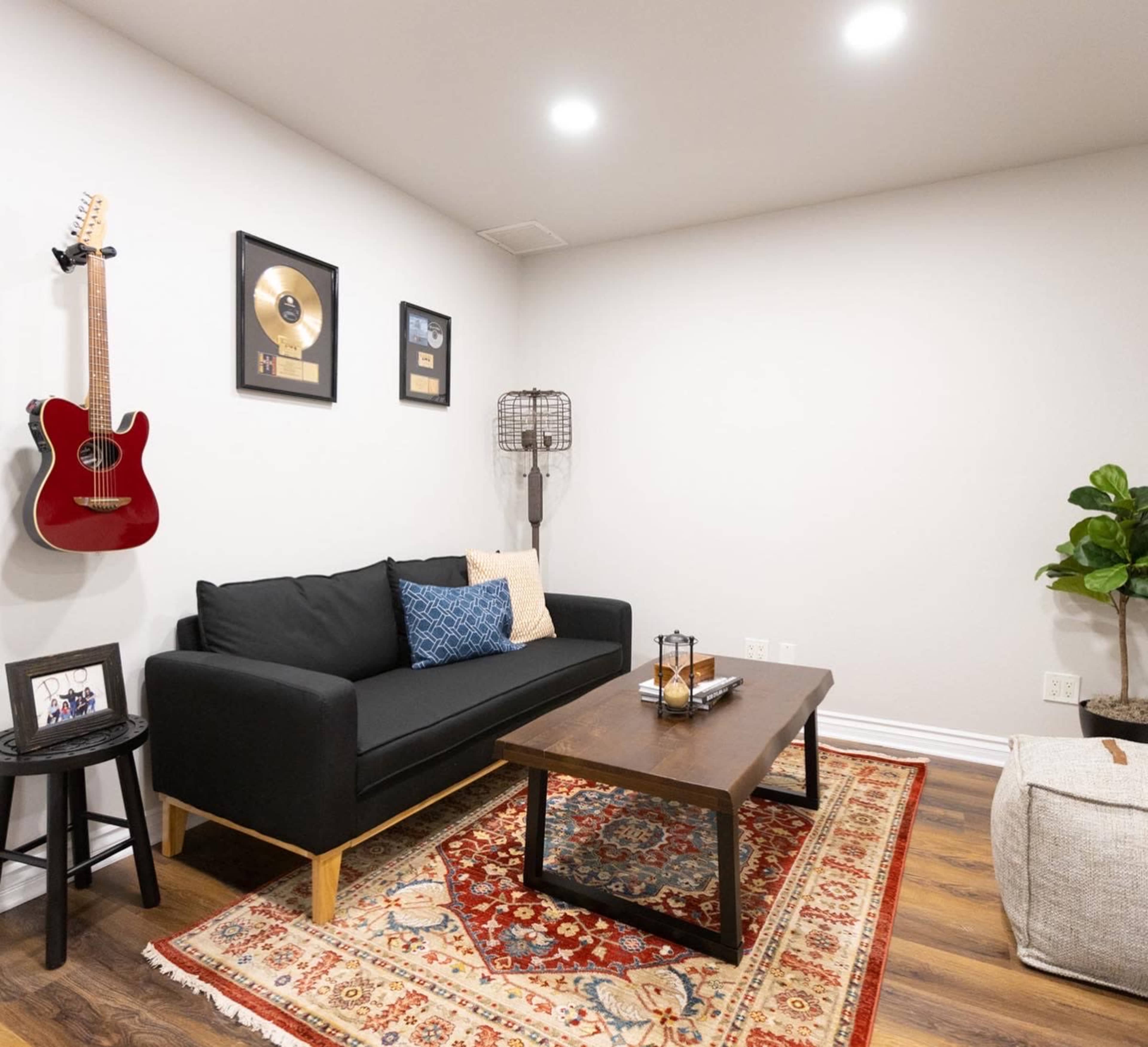 A modern living room features a black sofa, a wooden coffee table, and a decorative rug, with framed artwork on the walls and a guitar hanging nearby.