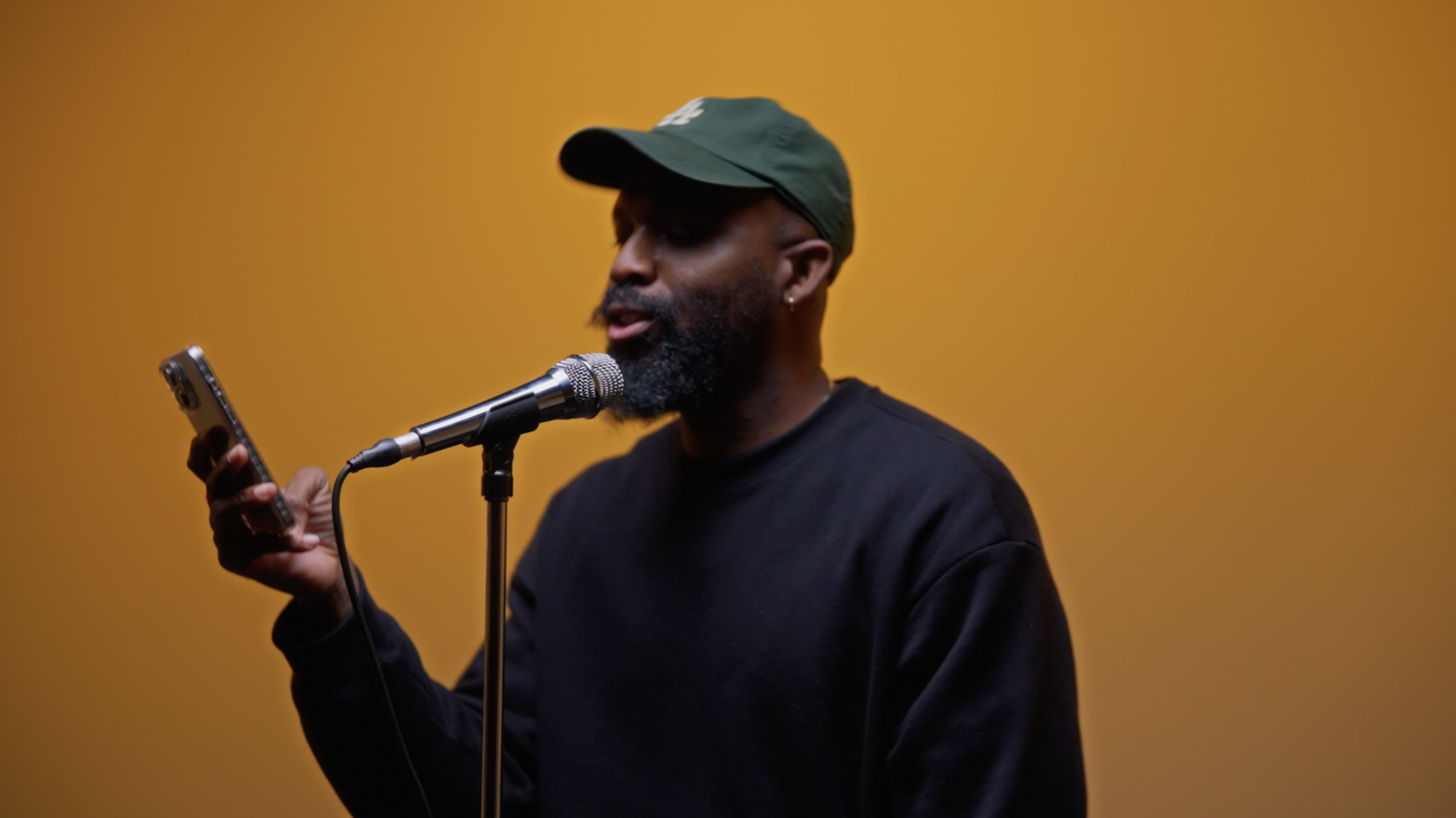 A man in a green cap holds a smartphone while speaking into a microphone against a bright yellow background.