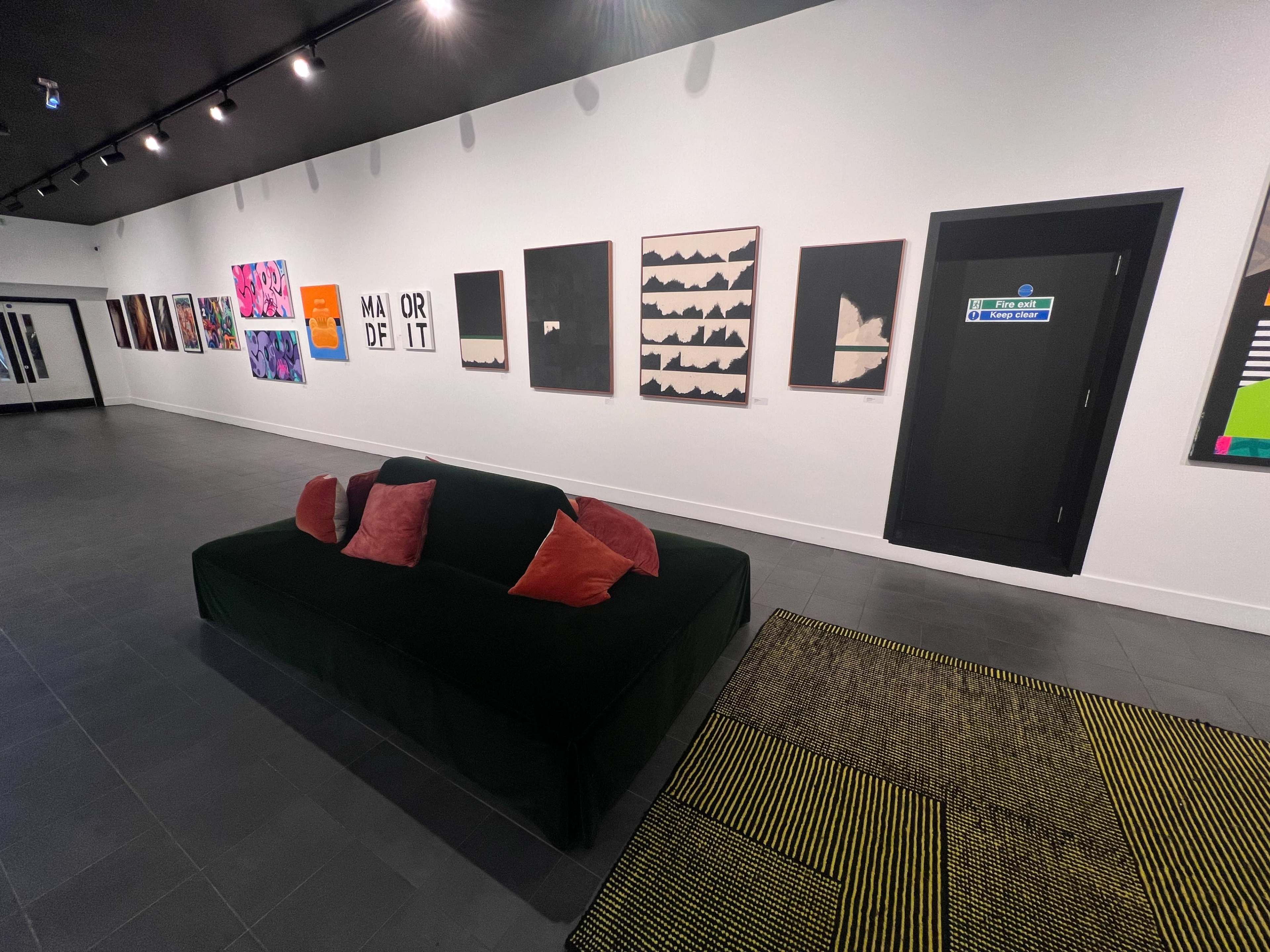 A contemporary art gallery featuring a green sofa with red and orange pillows, against a wall displaying various artworks and a door labeled with signage.