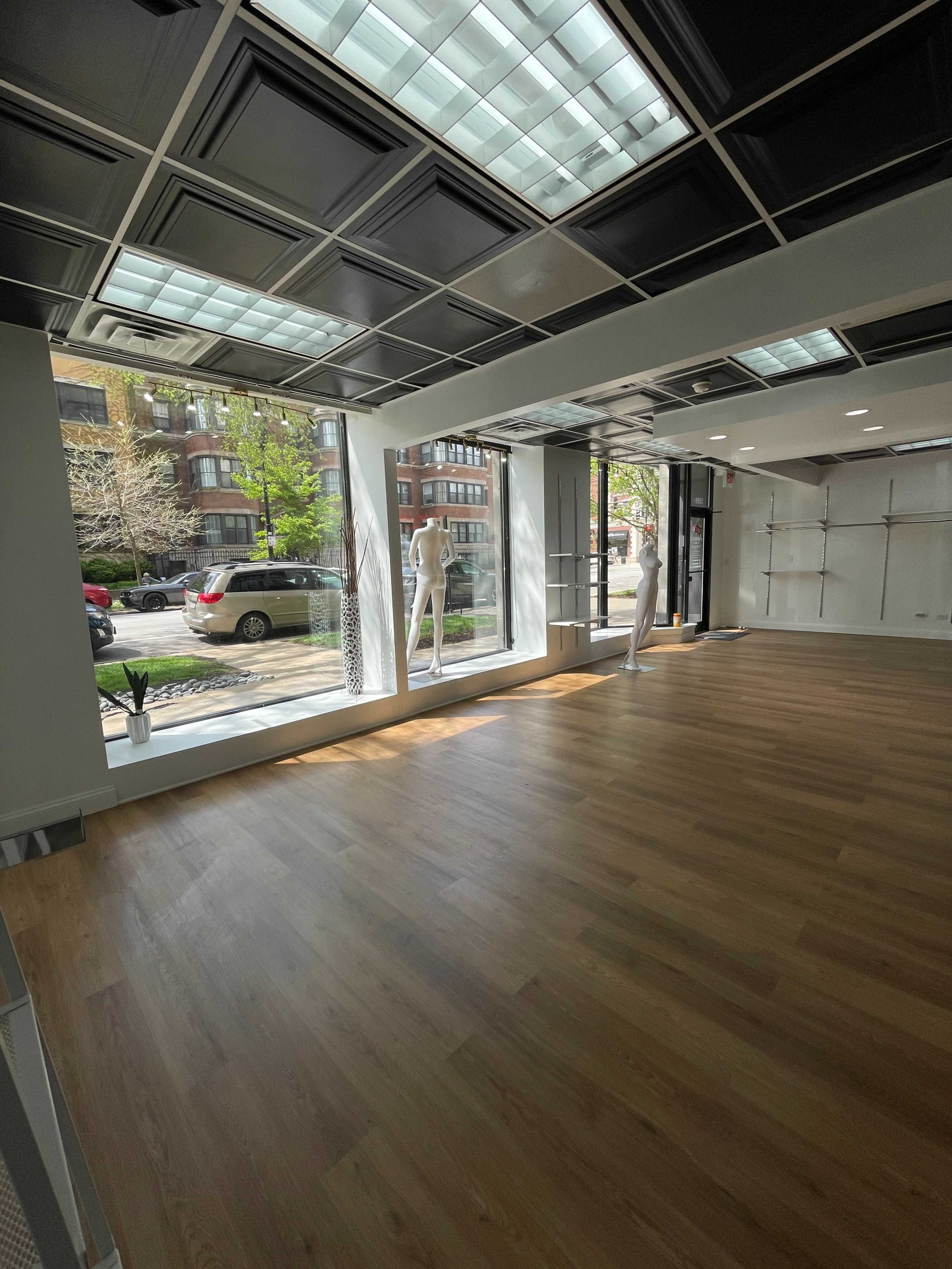 The image shows a spacious retail store interior with light wood flooring, large front windows, and two mannequins positioned by the windows.